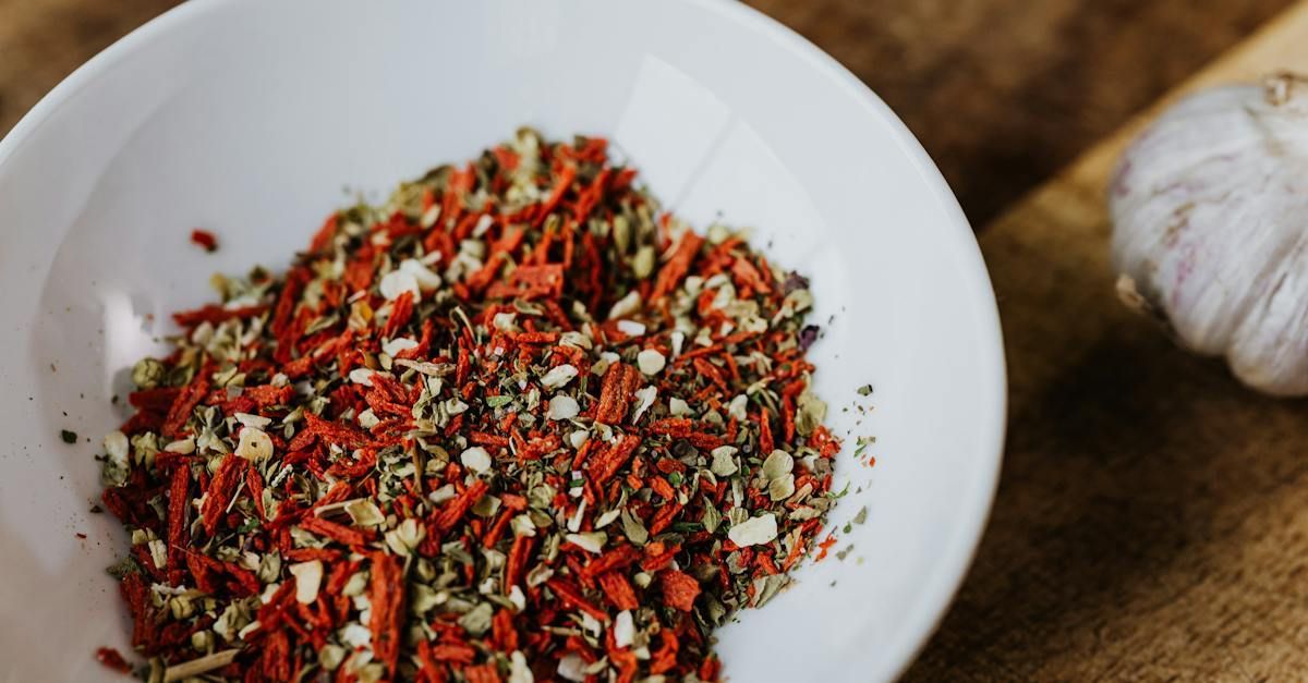 A bowl of colorful dried spice flakes on a wooden surface, with garlic cloves nearby