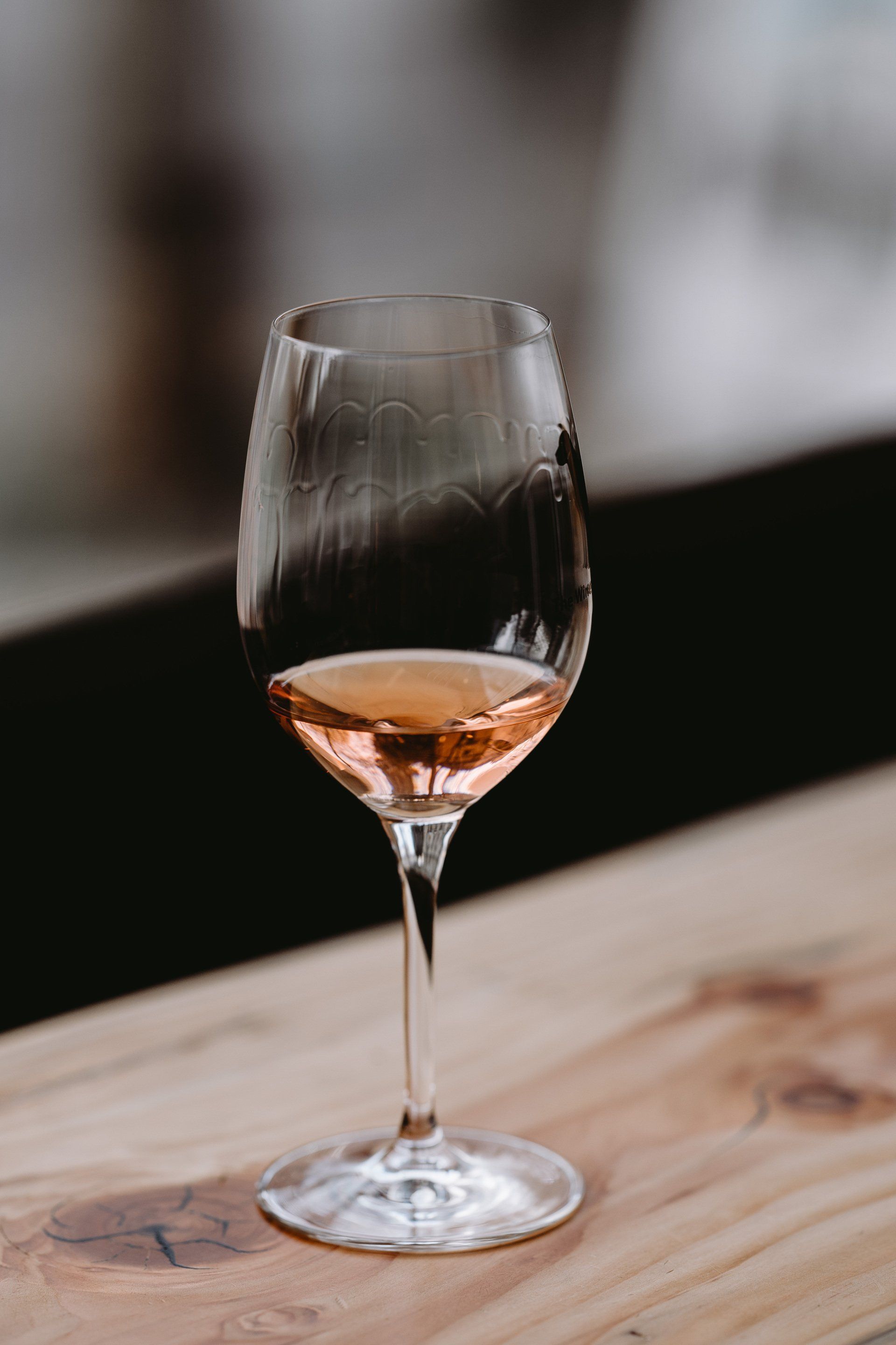 A wine glass with rose-colored liquid sits on a wooden surface. The background is blurred.