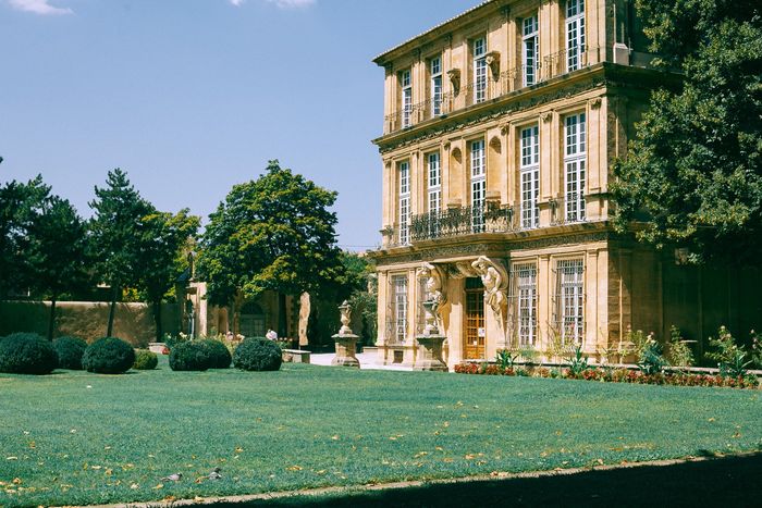 Three-story beige building with ornate facade, in a sunny green yard with trees.
