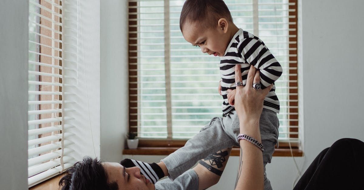 Person lying down lifting a baby wearing striped shirt, by a window with blinds.
