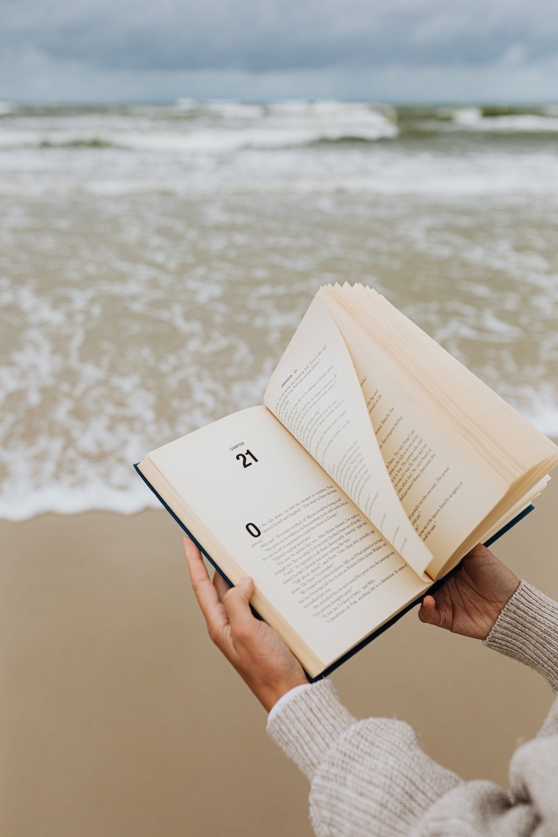 A person in a beige sweater holds an open book with page 21 visible, with the ocean waves in the background.
