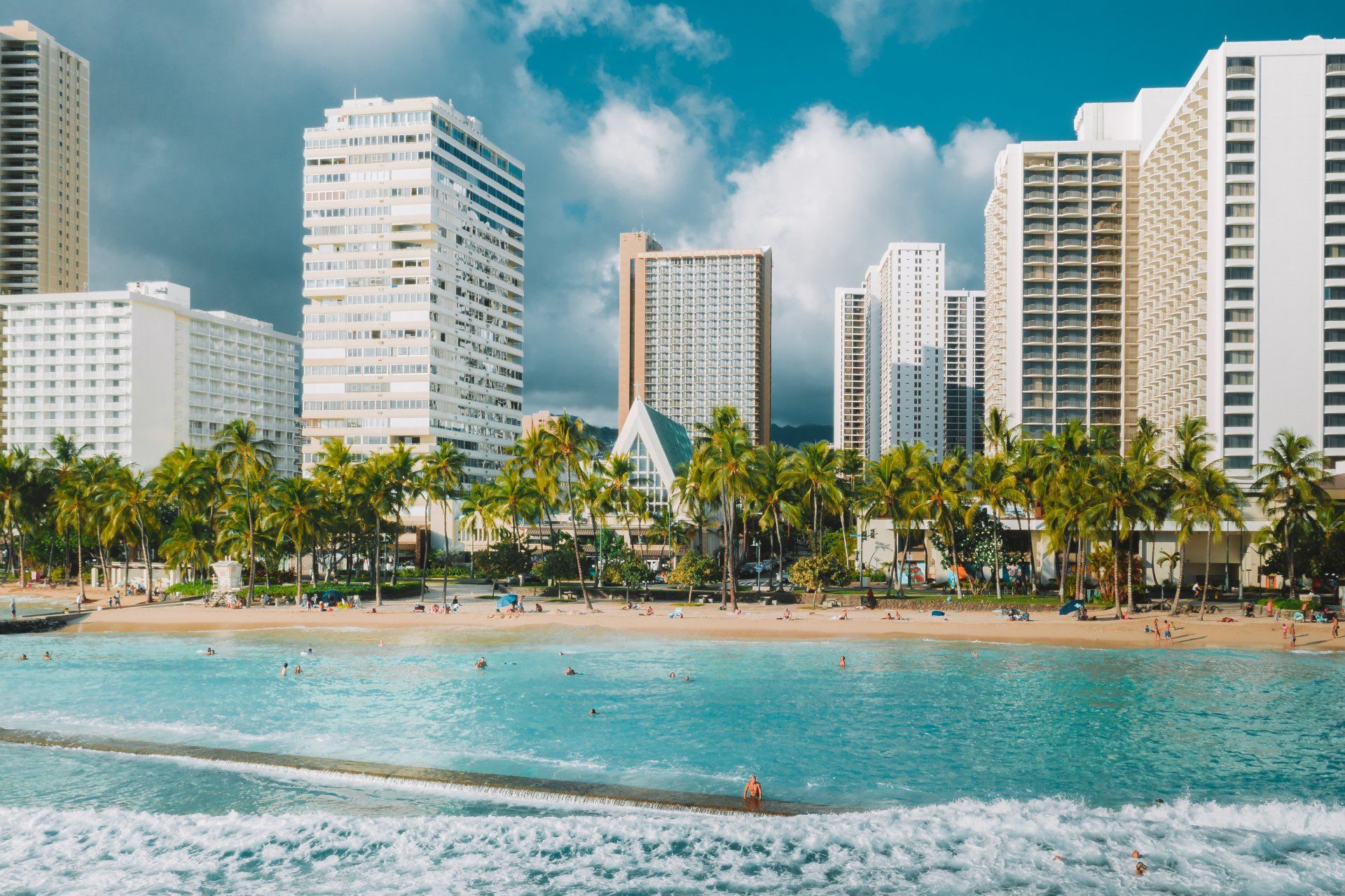 A sunny beach scene in Honolulu, Hawaii, featuring high-rise resort hotels, palm trees, and turquoise ocean waves.