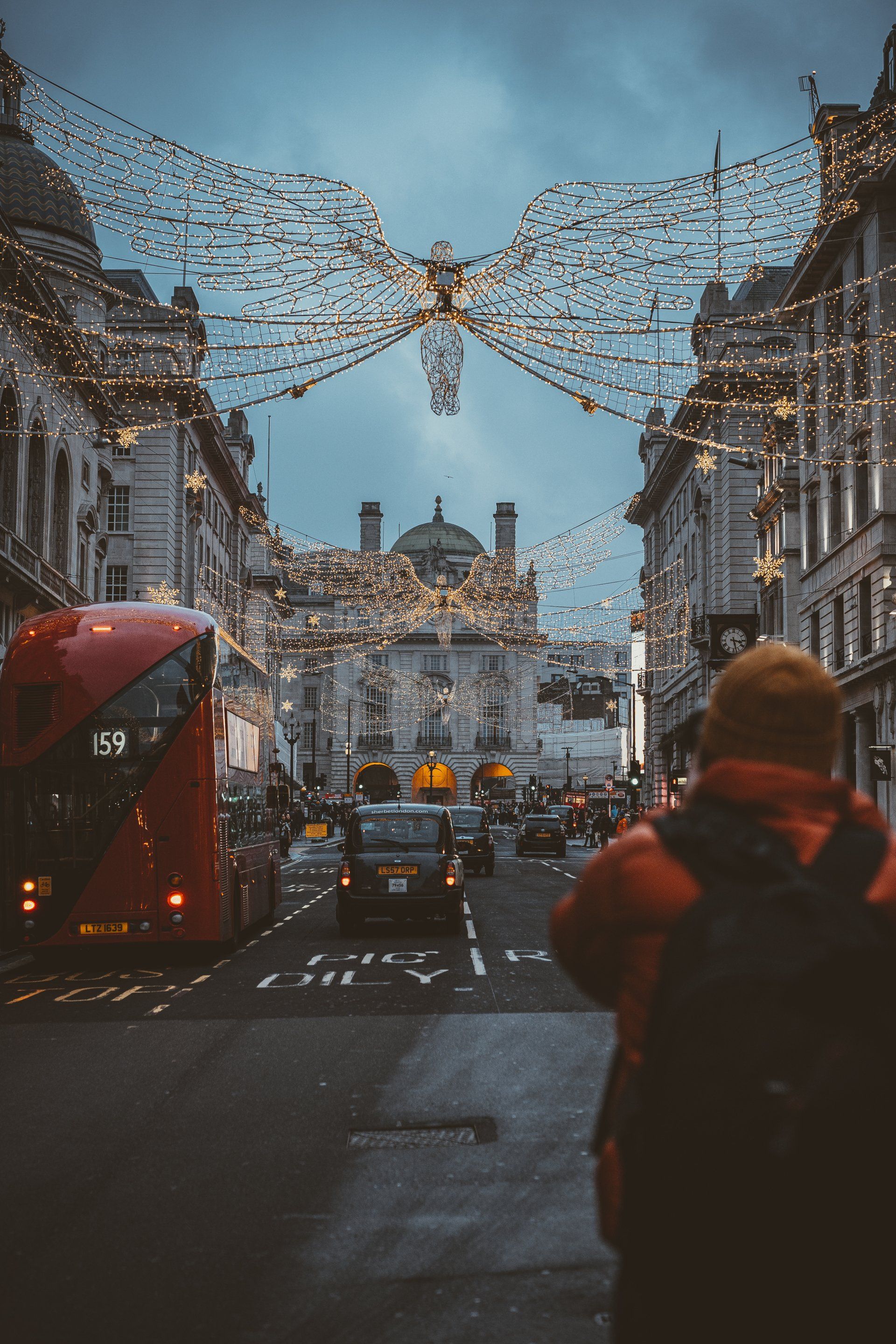 London street with Christmas lights. Red bus, person with backpack, cars, and buildings under the illuminated decorations.