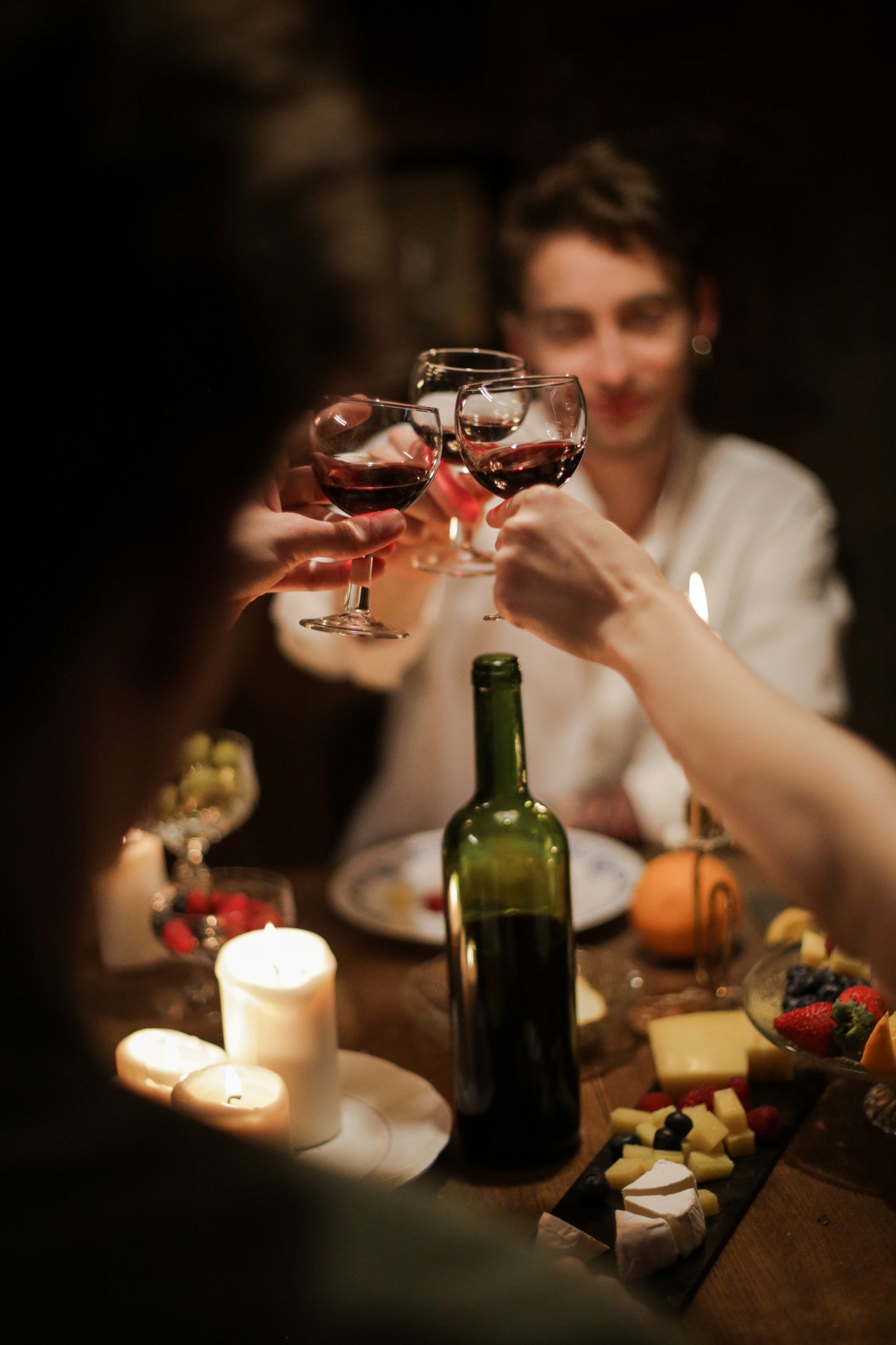 People toasting with red wine glasses at a dimly lit table with food, candles, and a wine bottle.