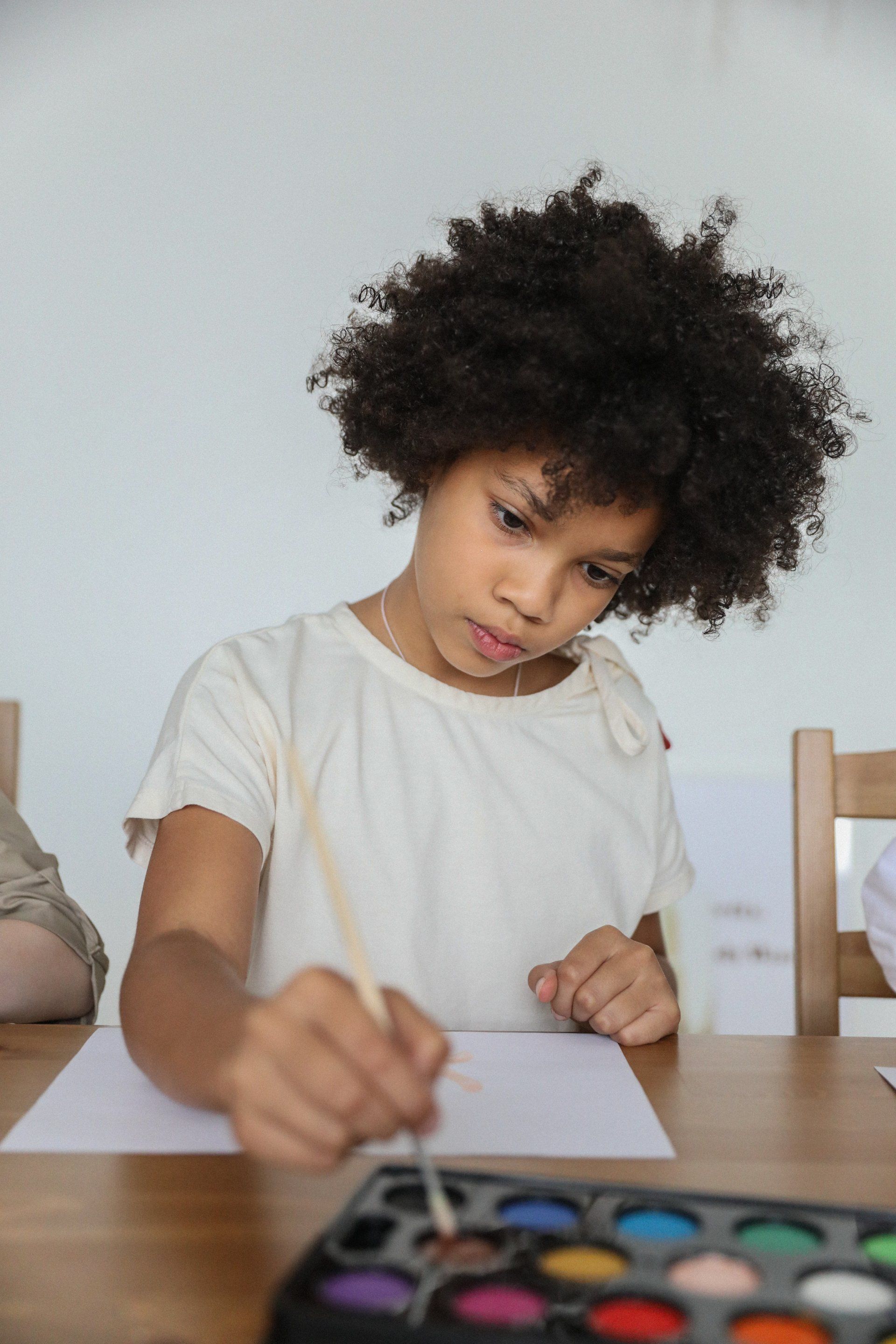 Girl with afro painting at a table with watercolor paints, focused expression.
