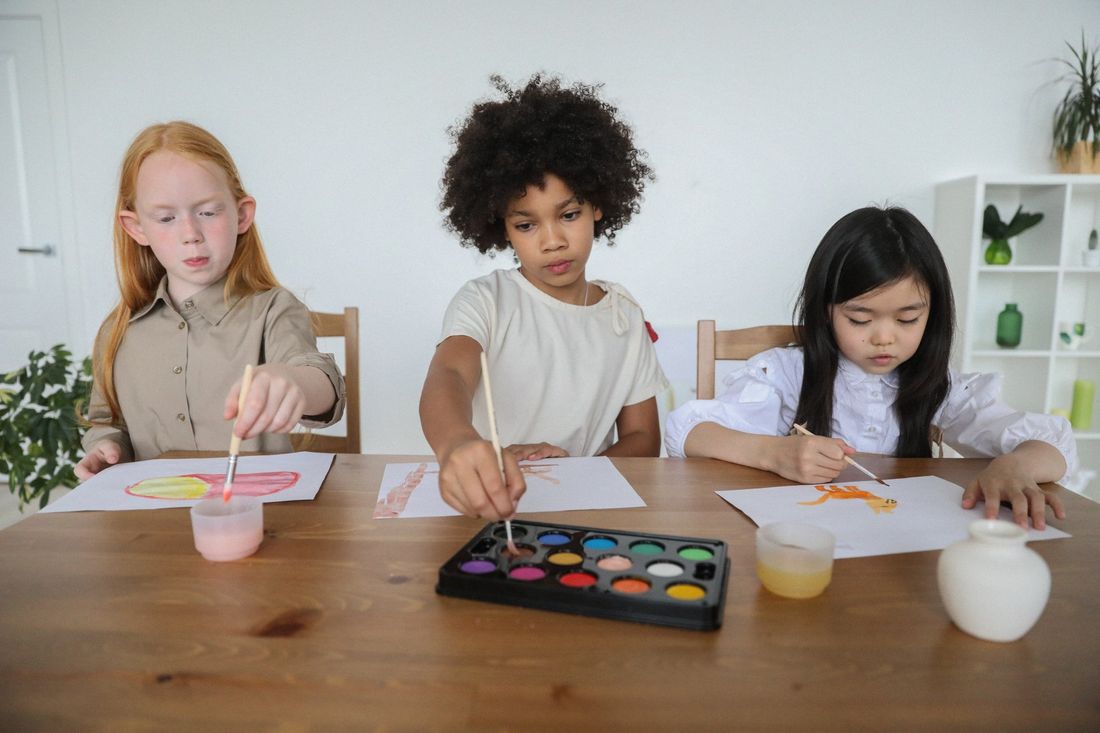 Three children are sitting at a table painting with watercolors.