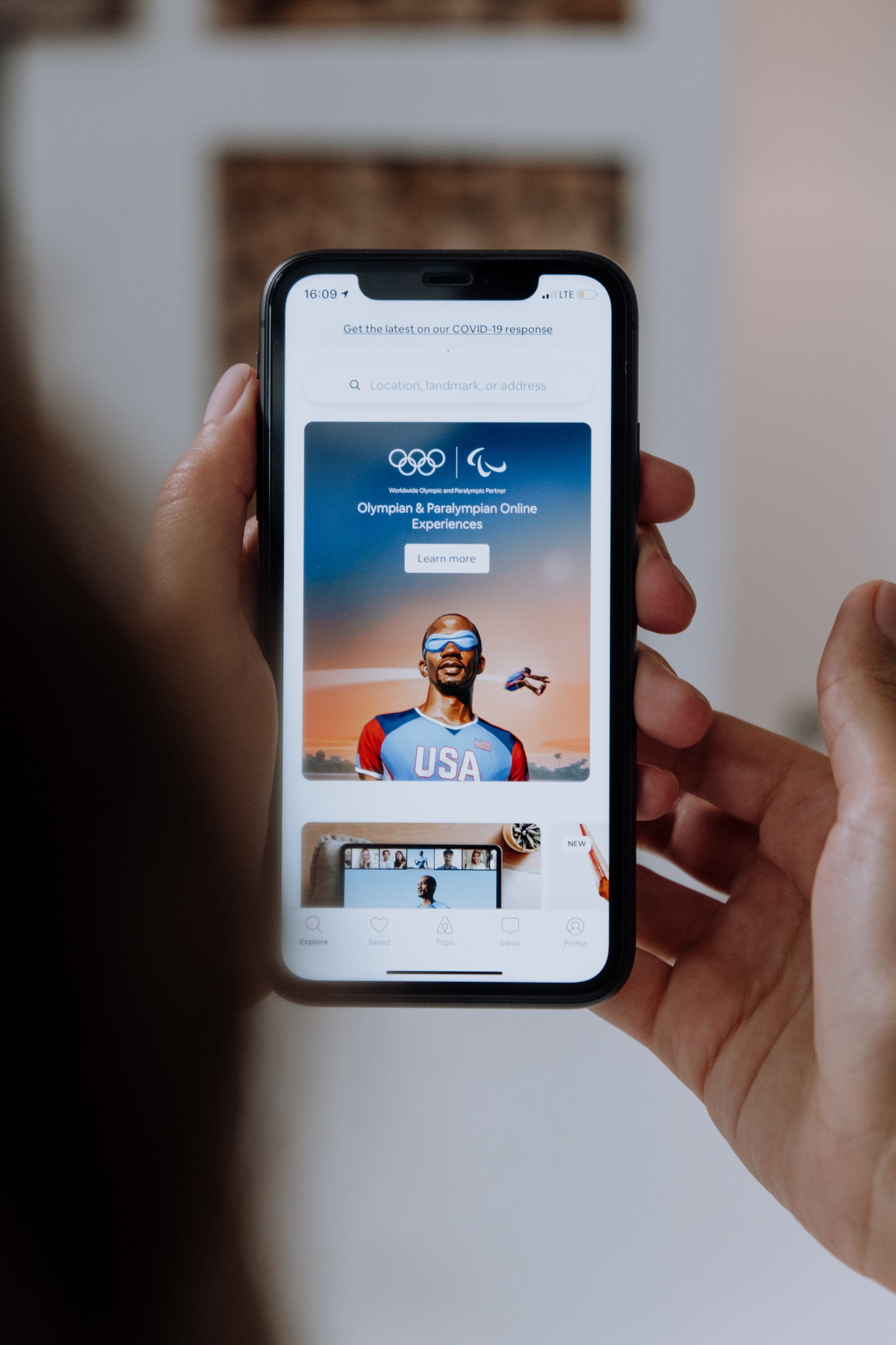 Mãos segurando um smartphone exibindo o site dos Jogos Olímpicos e Paralímpicos com a foto de um atleta dos EUA.