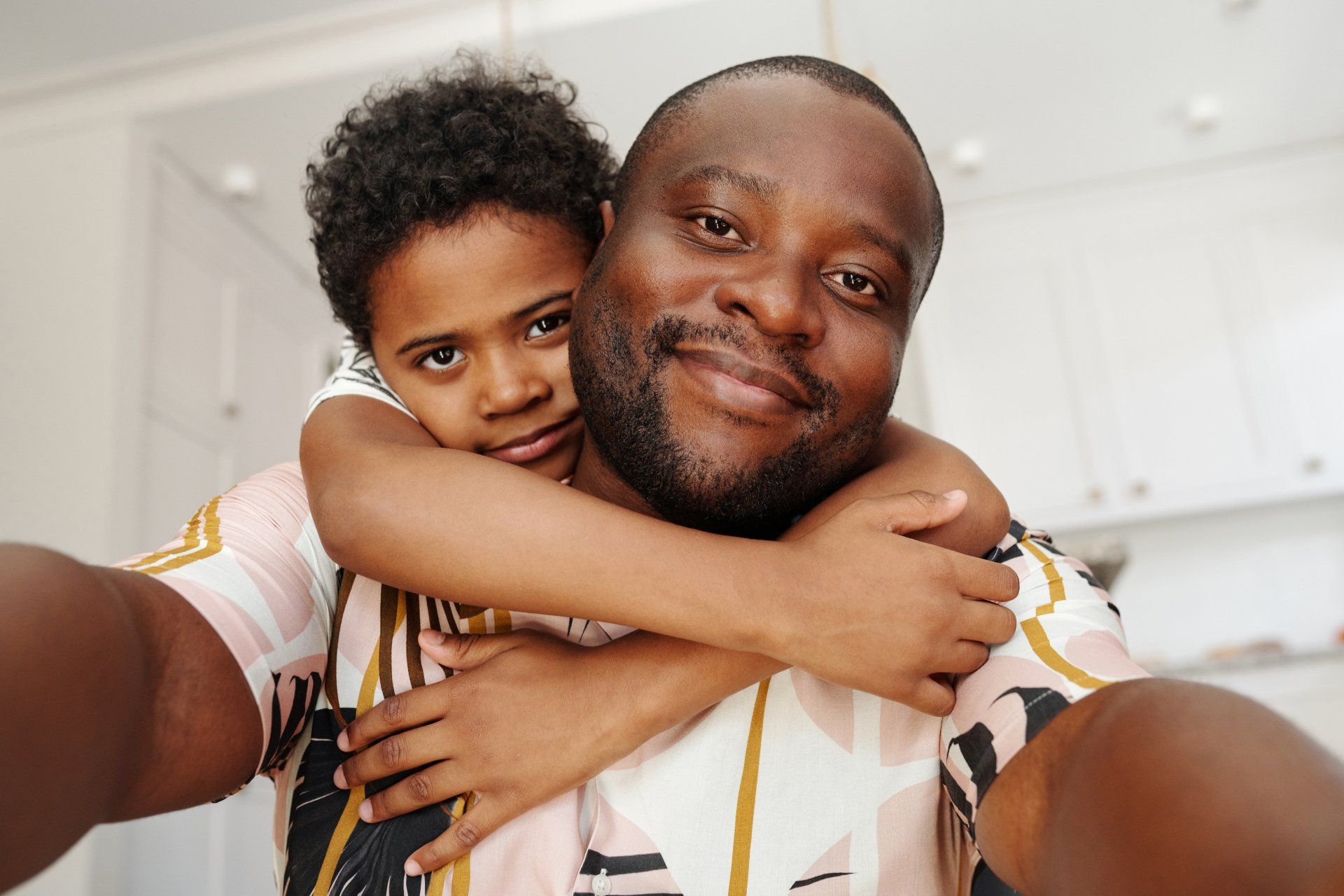 A person takes a selfie with a child hugging from behind. Both smiling. Indoors, neutral colors.