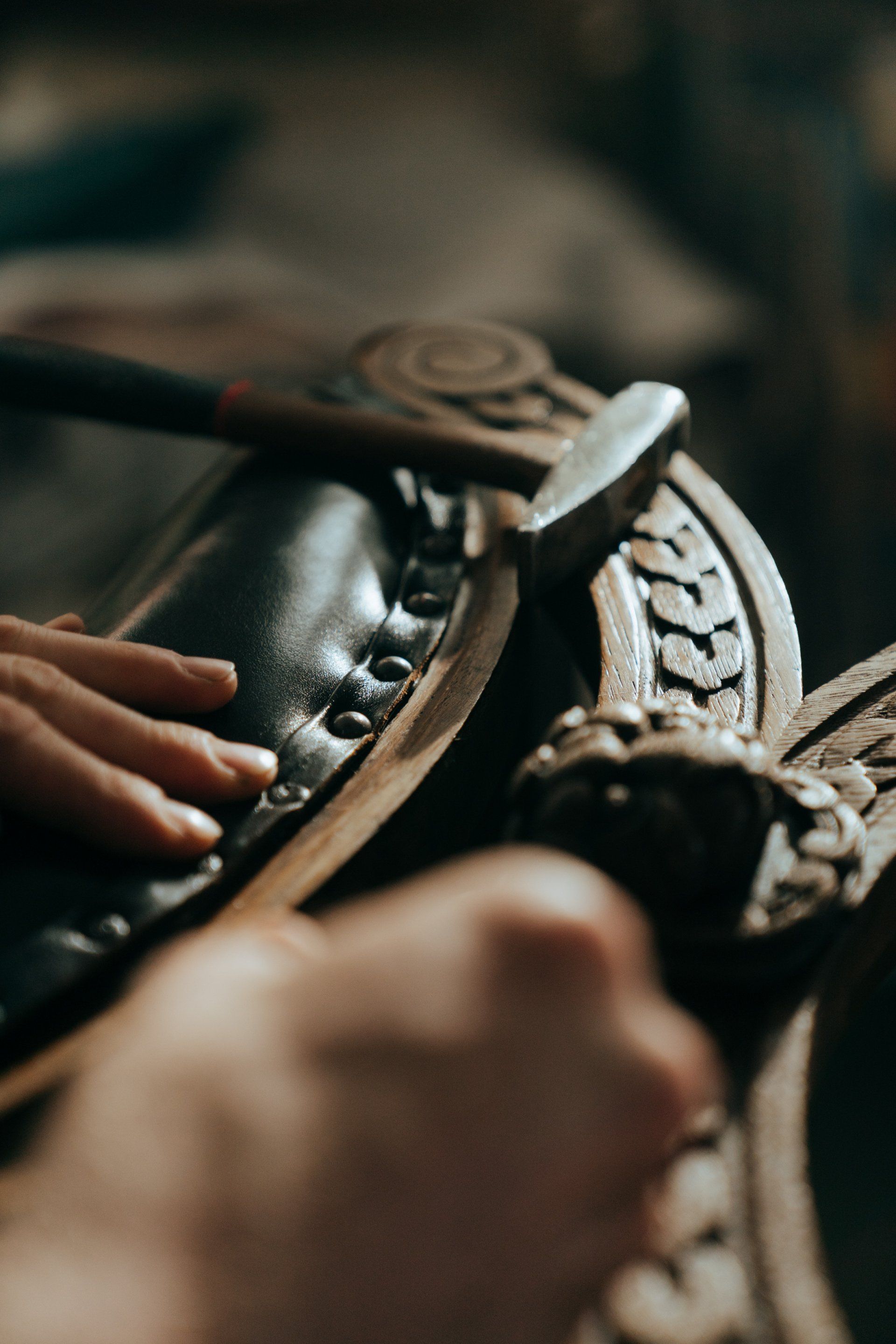 Des mains façonnent une lanière de cuir ornée à l'aide d'un marteau et d'outils sur un établi.