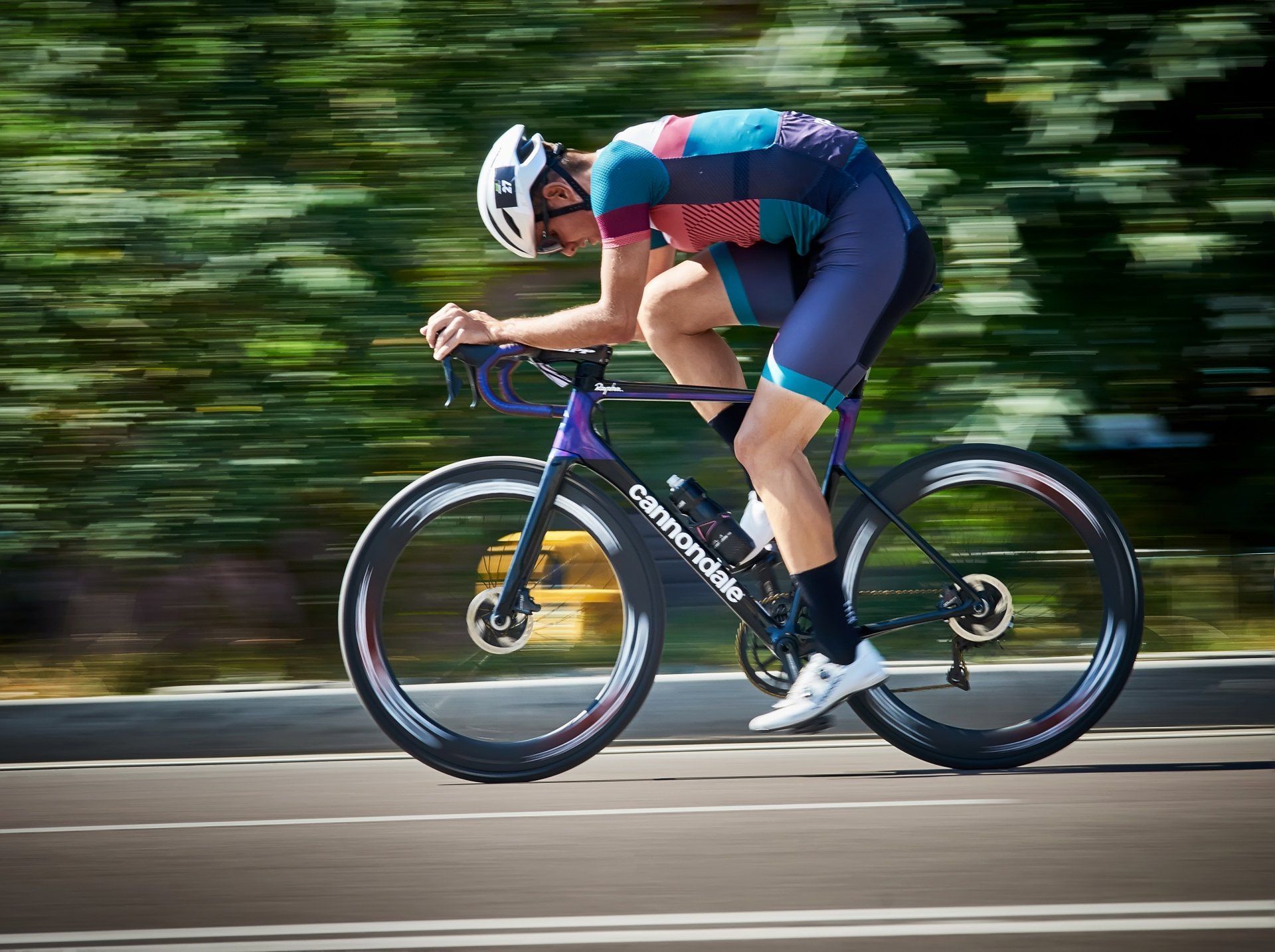 Cyclist in aerodynamic position on road bike, wearing helmet and colorful gear, blurred background.