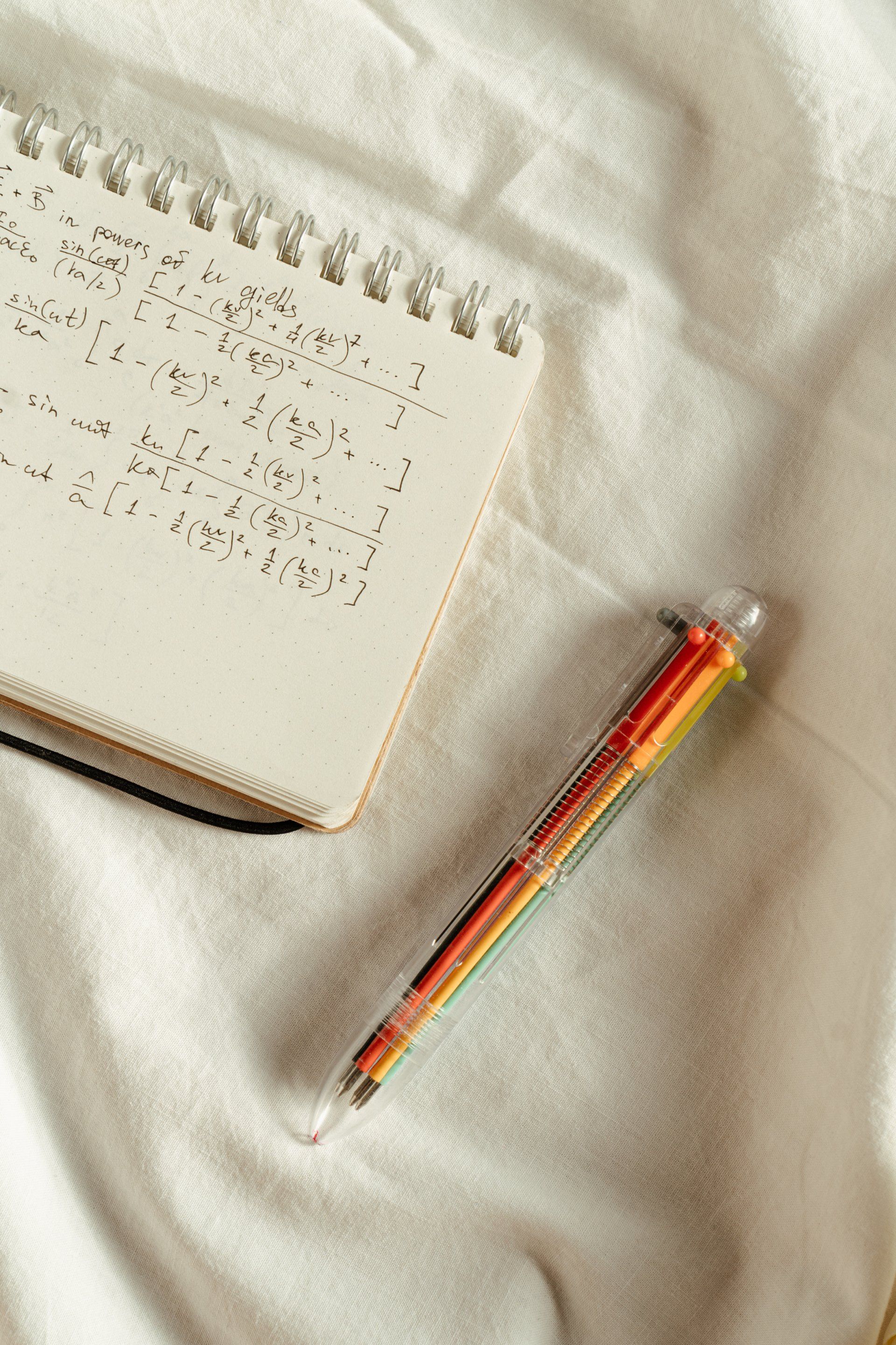 A rainbow pen is sitting next to a notebook on a bed.
