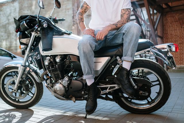 Person with tattoos sits on a white motorcycle, wearing jeans and boots, outdoors.