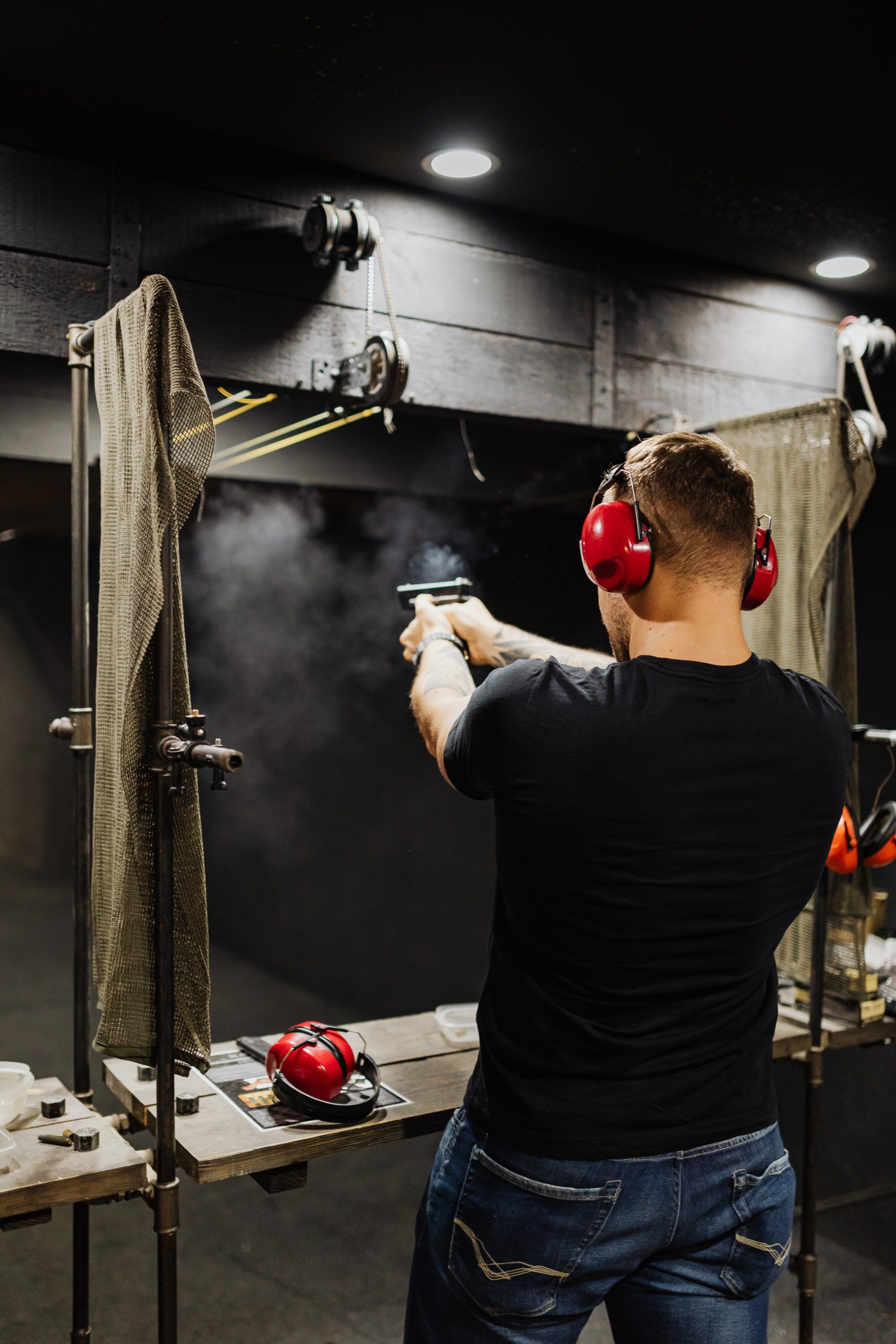 Person shooting a handgun at an indoor firing range, wearing red earmuffs and a black shirt.