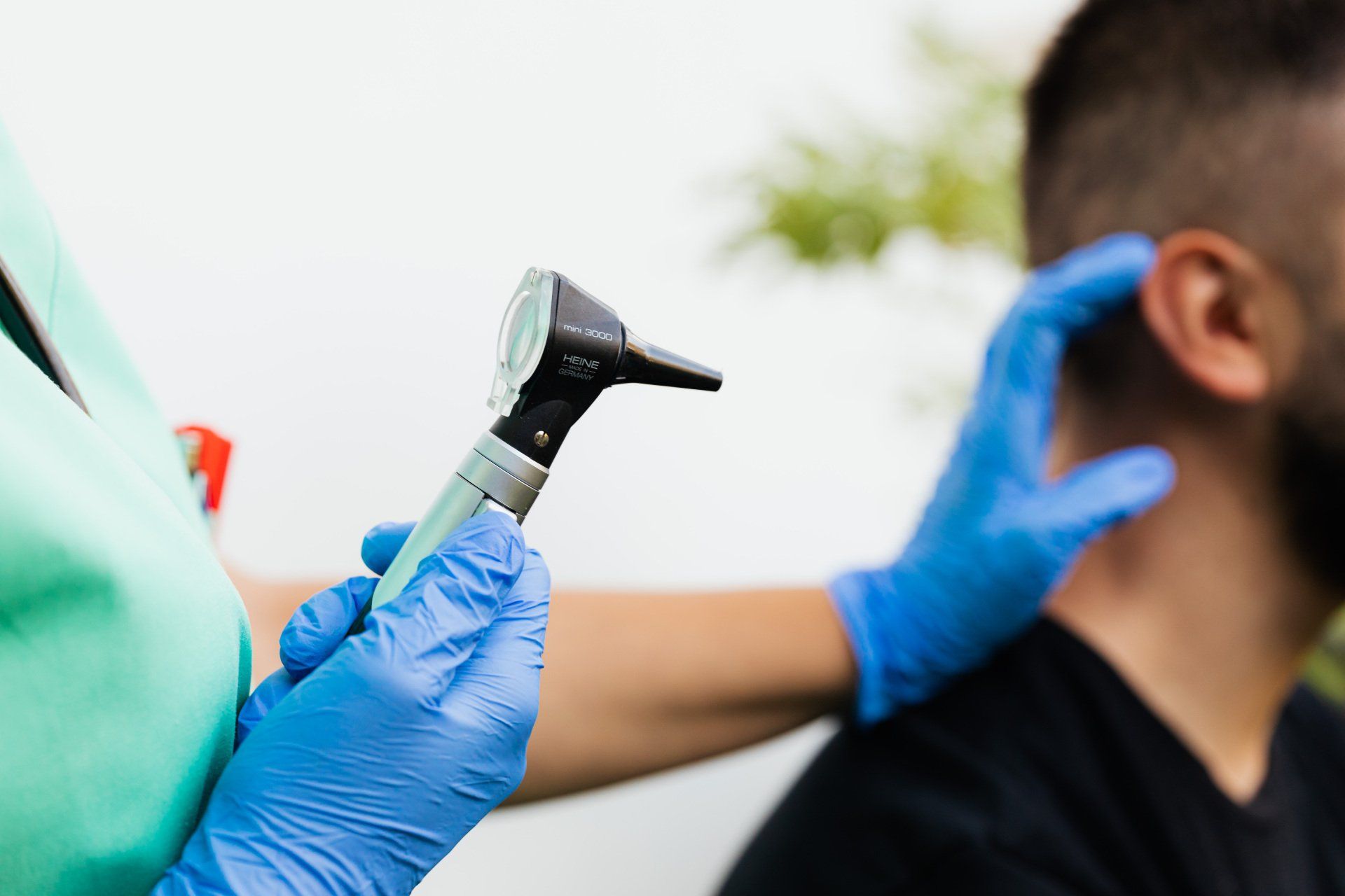 Healthcare worker using otoscope to examine a seated patient’s ear, wearing blue gloves.