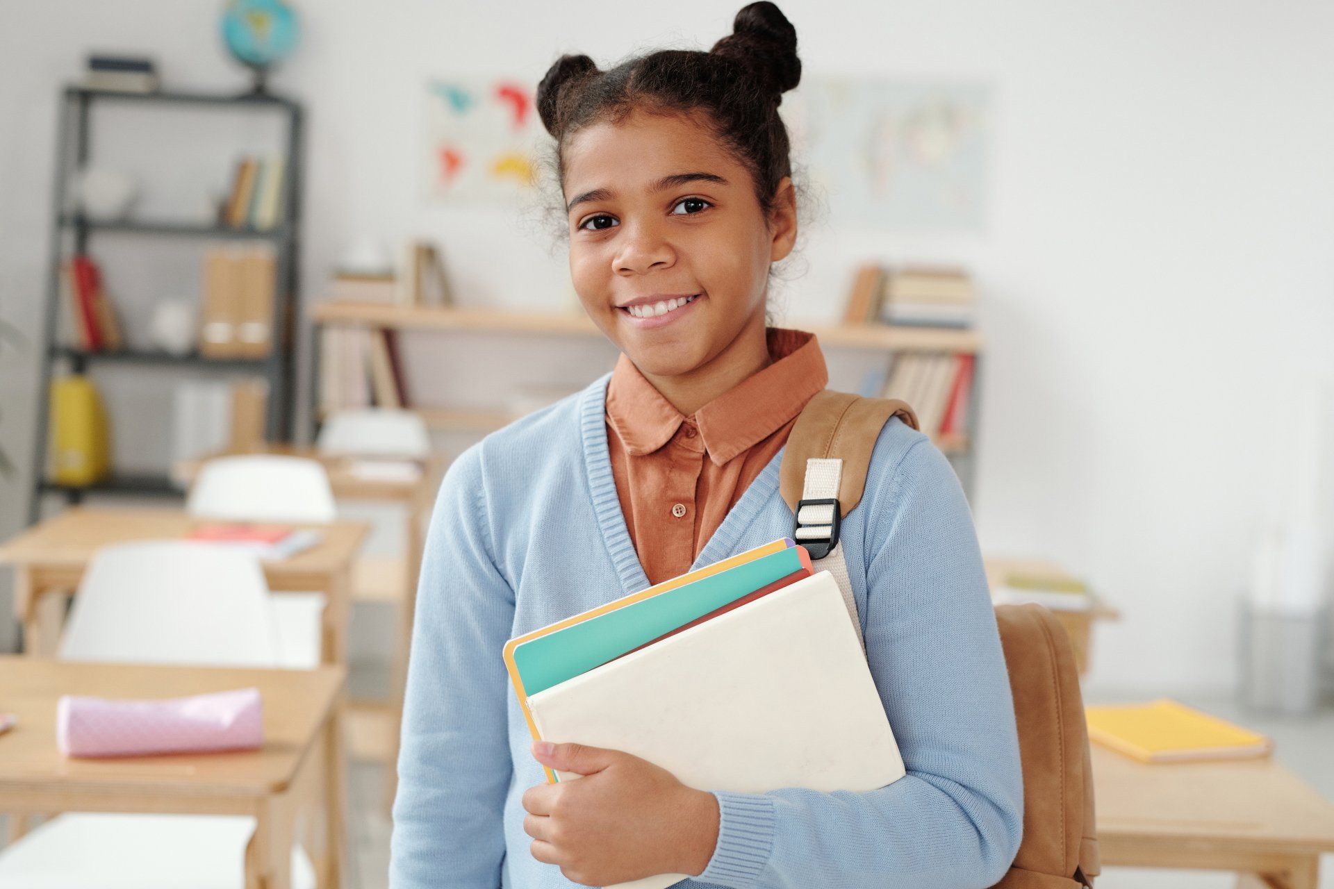 A young girl is holding books and a backpack in a classroom.