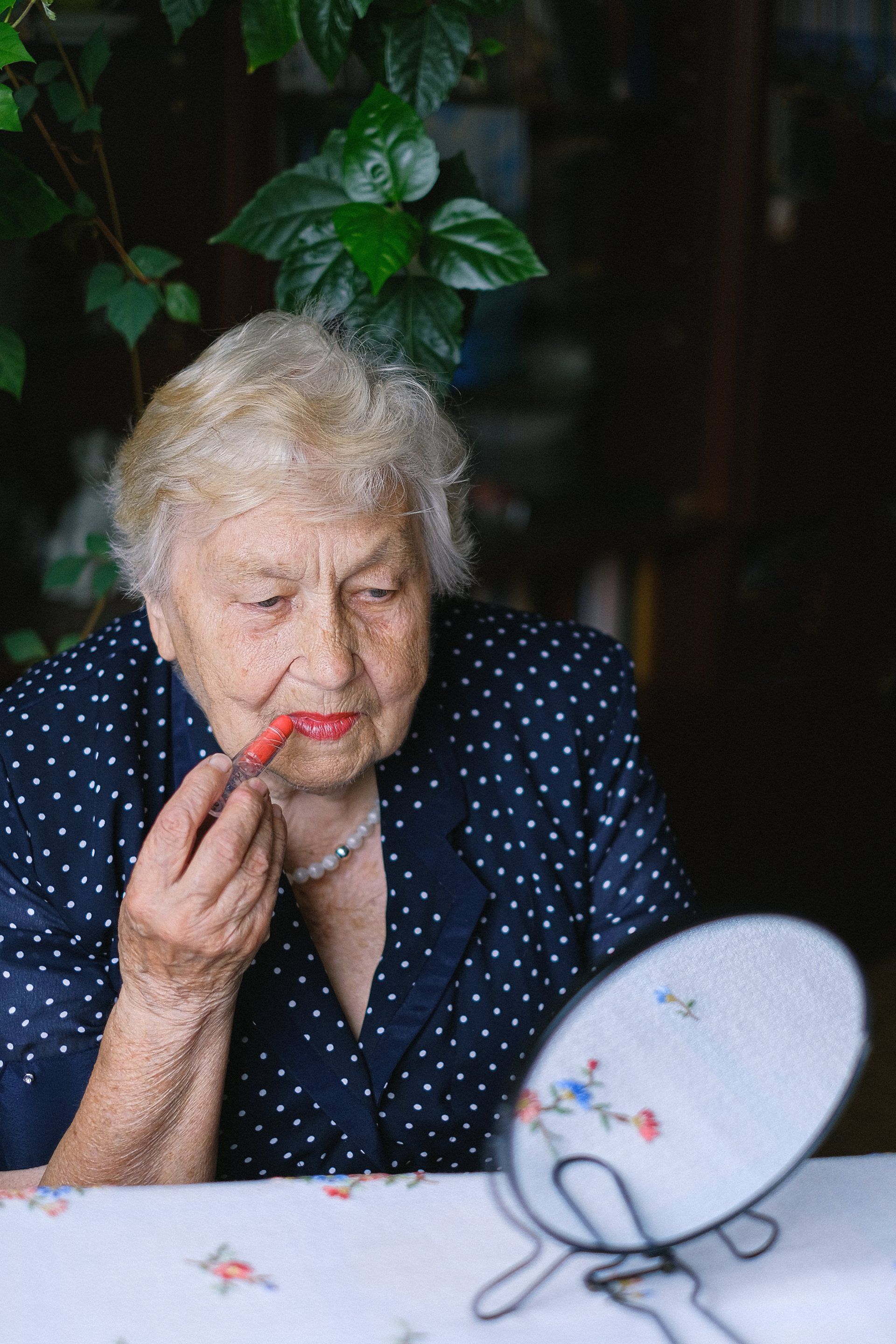 Elderly woman applying red lipstick, looking in a small hand mirror.