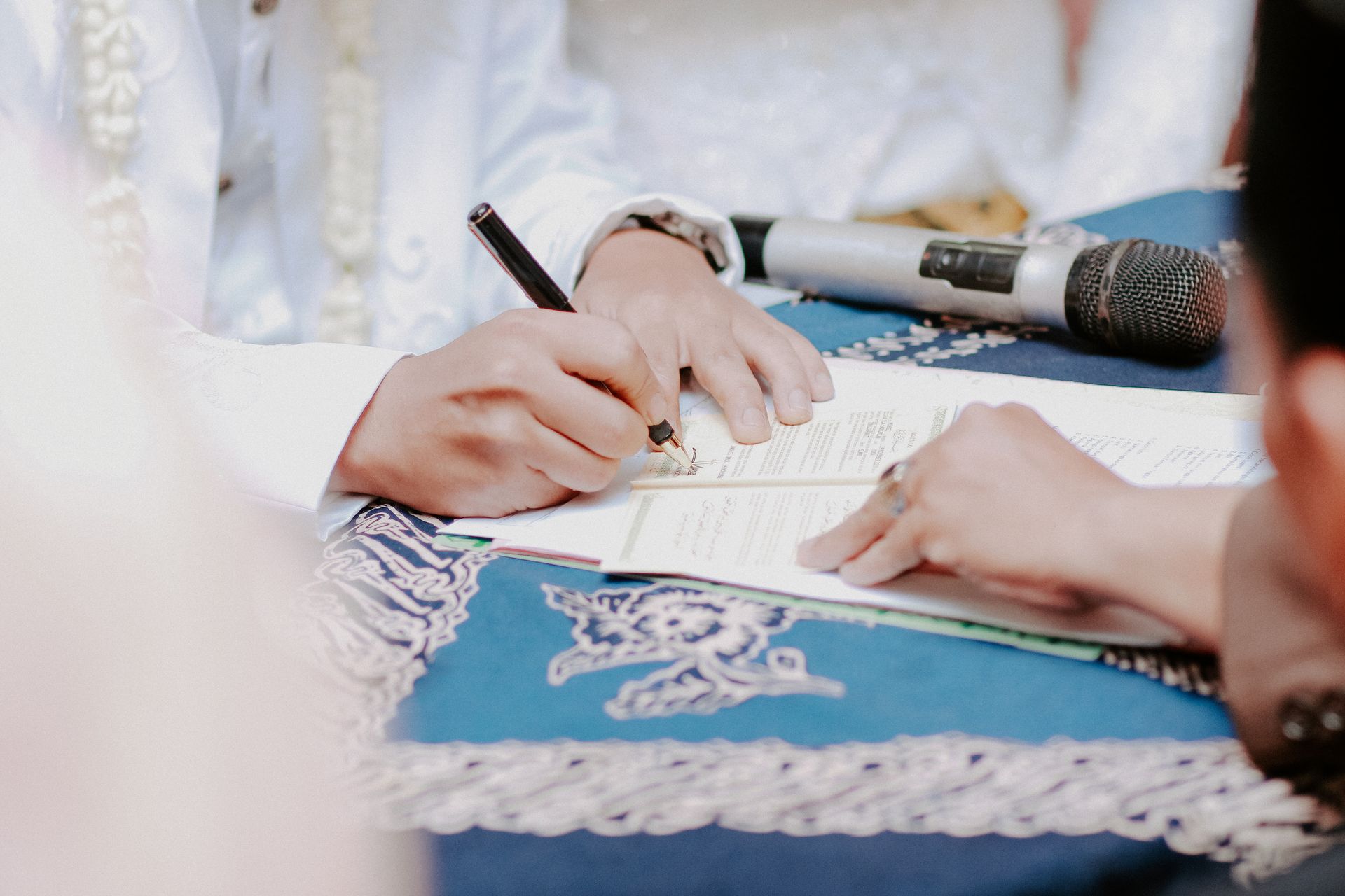 Des mains écrivent sur un document posé sur une nappe bleue à motifs, un microphone se trouvant à proximité.