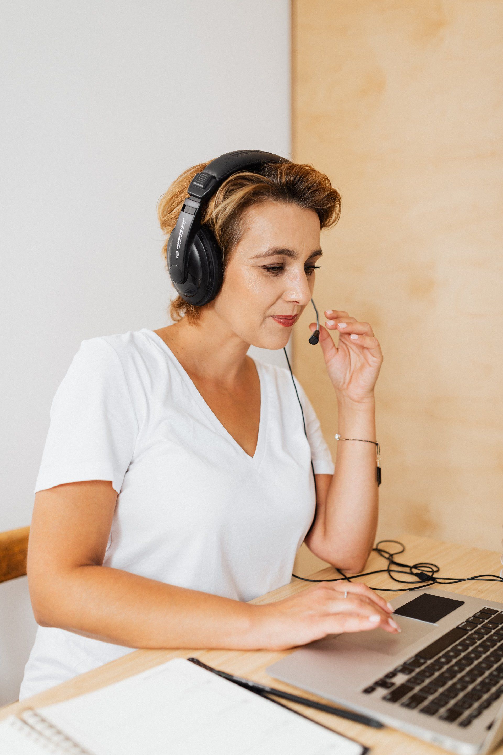 Person wearing headphones and a white shirt works on a laptop at a desk, speaking into a microphone.