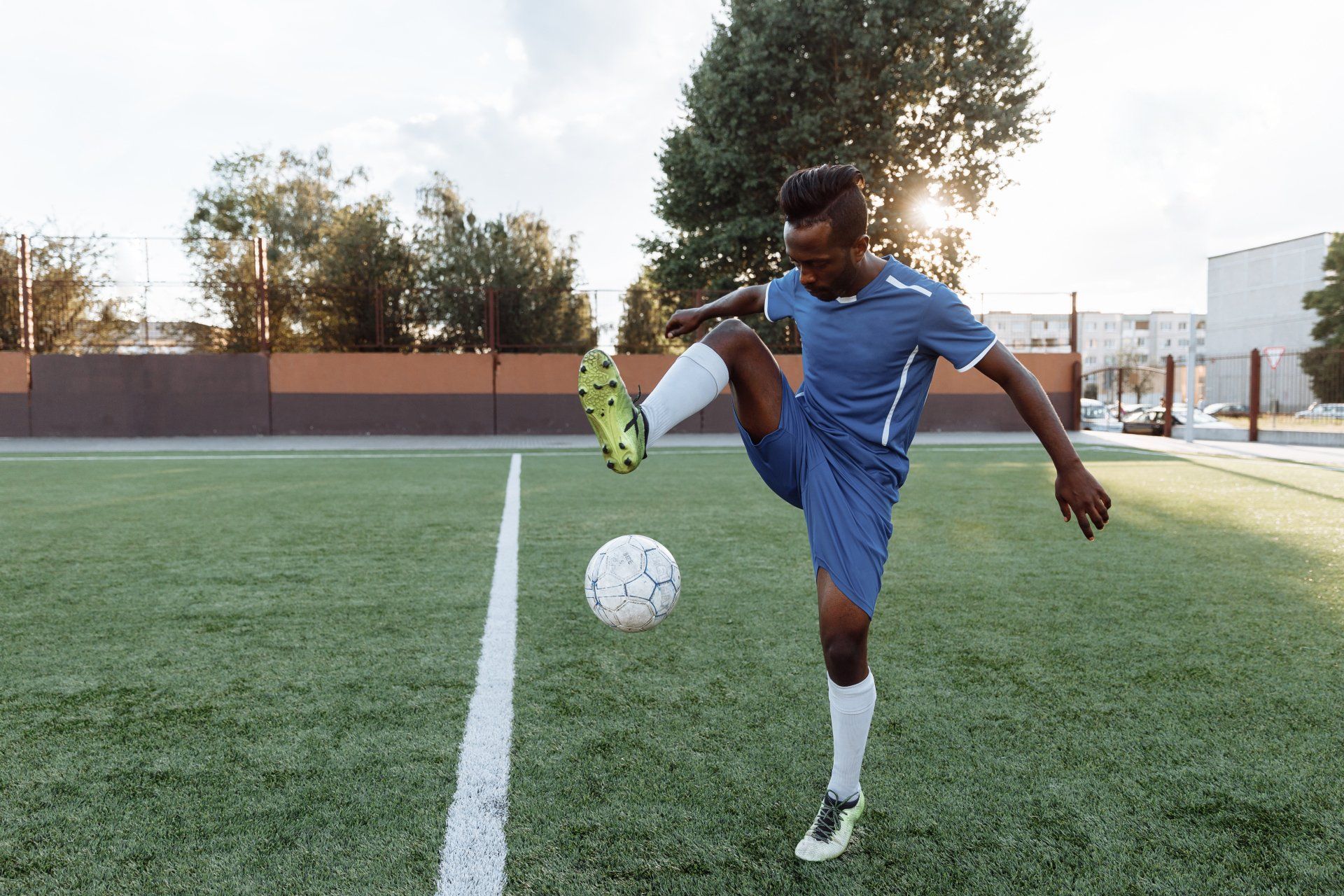 Soccer player in blue uniform kicking a ball on a grassy field at sunset