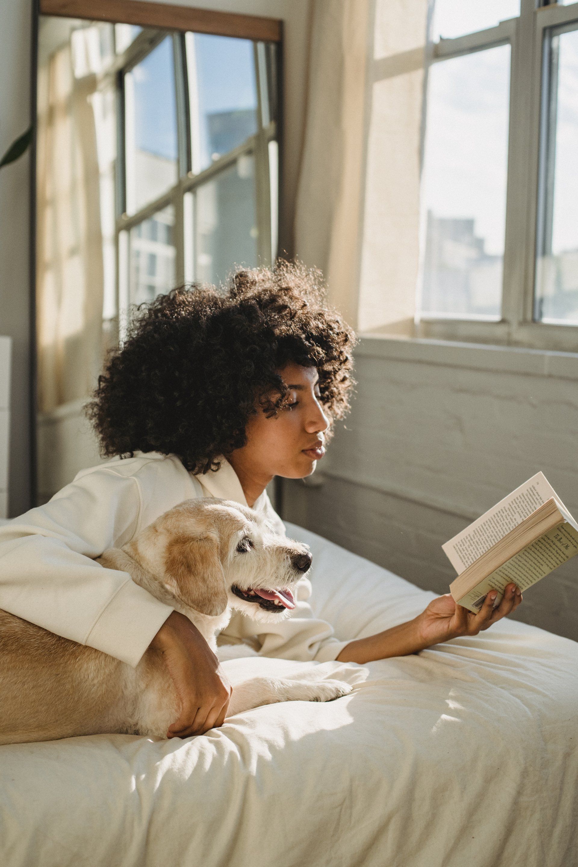 A woman is laying on a bed reading a book to her dog.