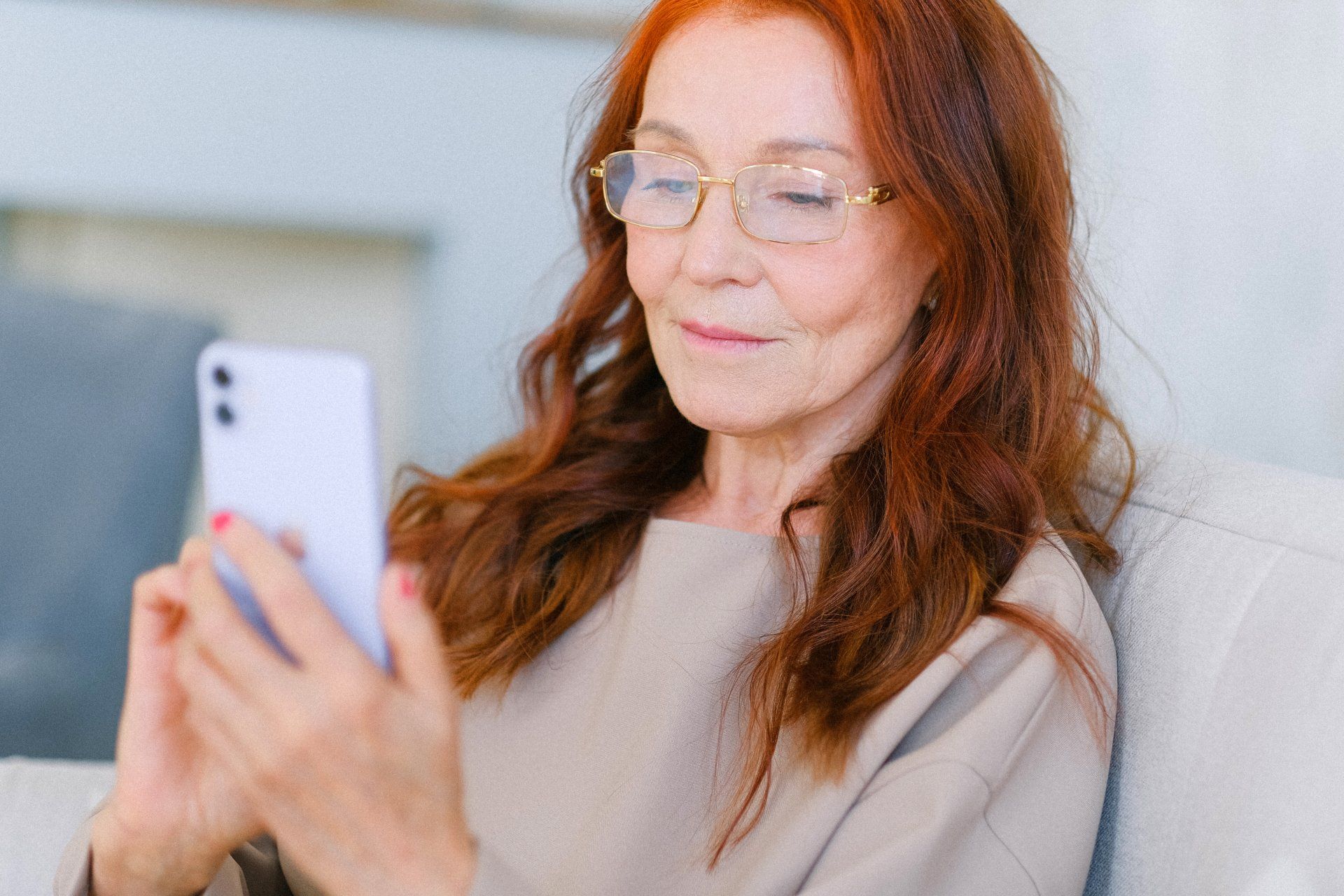 Woman with red hair wearing glasses, smiling at a smartphone on a couch.