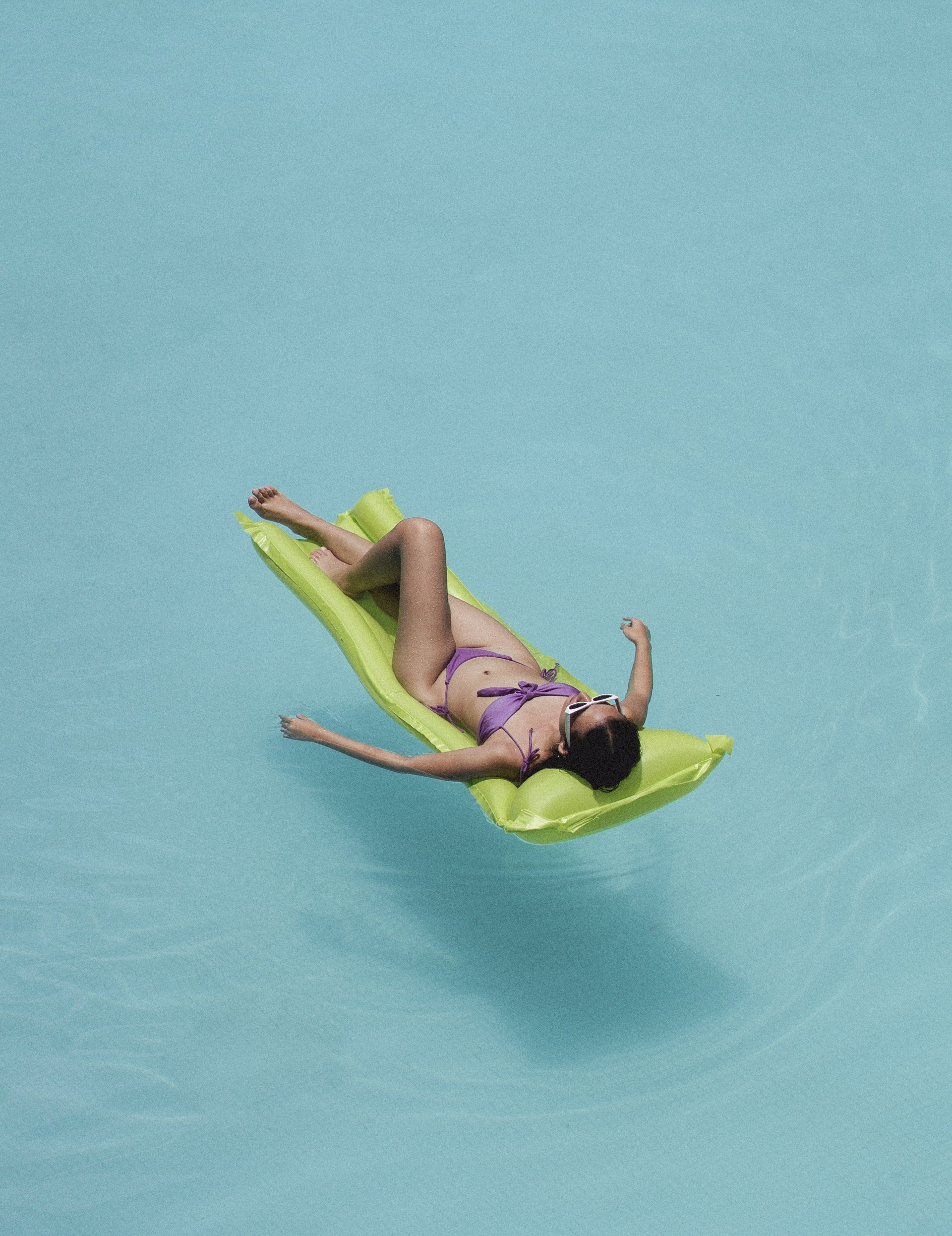 A woman in a bikini is floating on a green raft in a pool.