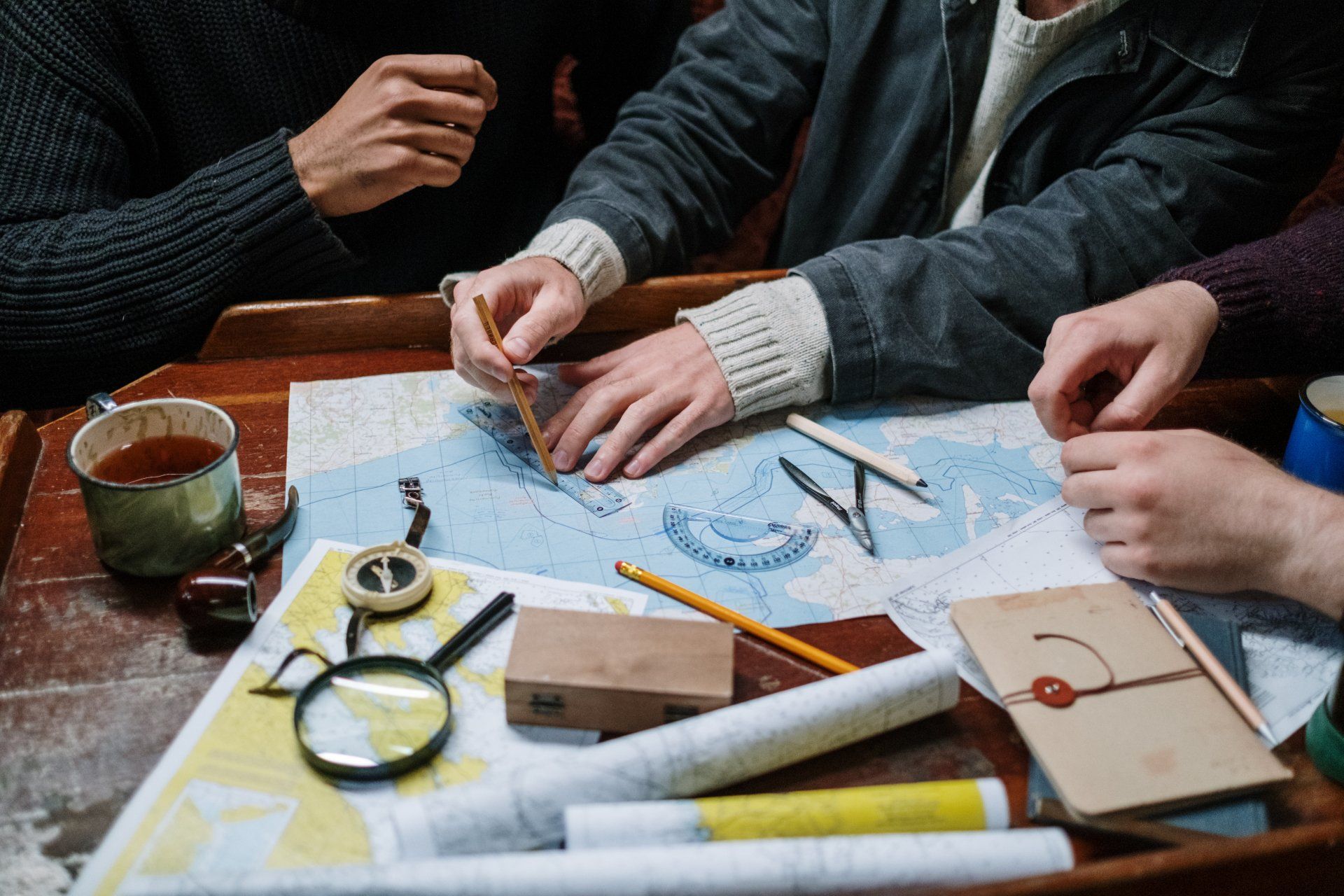 Drie mensen stippelen een route uit op een kaart. Diverse navigatiemiddelen liggen op een houten tafel.