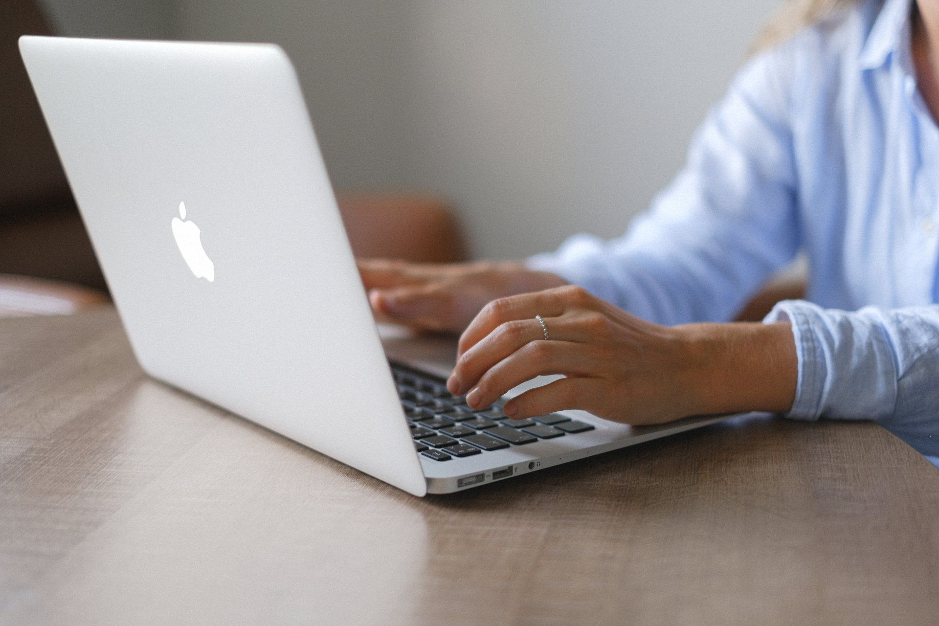 Person typing on a silver Macbook laptop on a wooden table, wearing a light blue shirt.