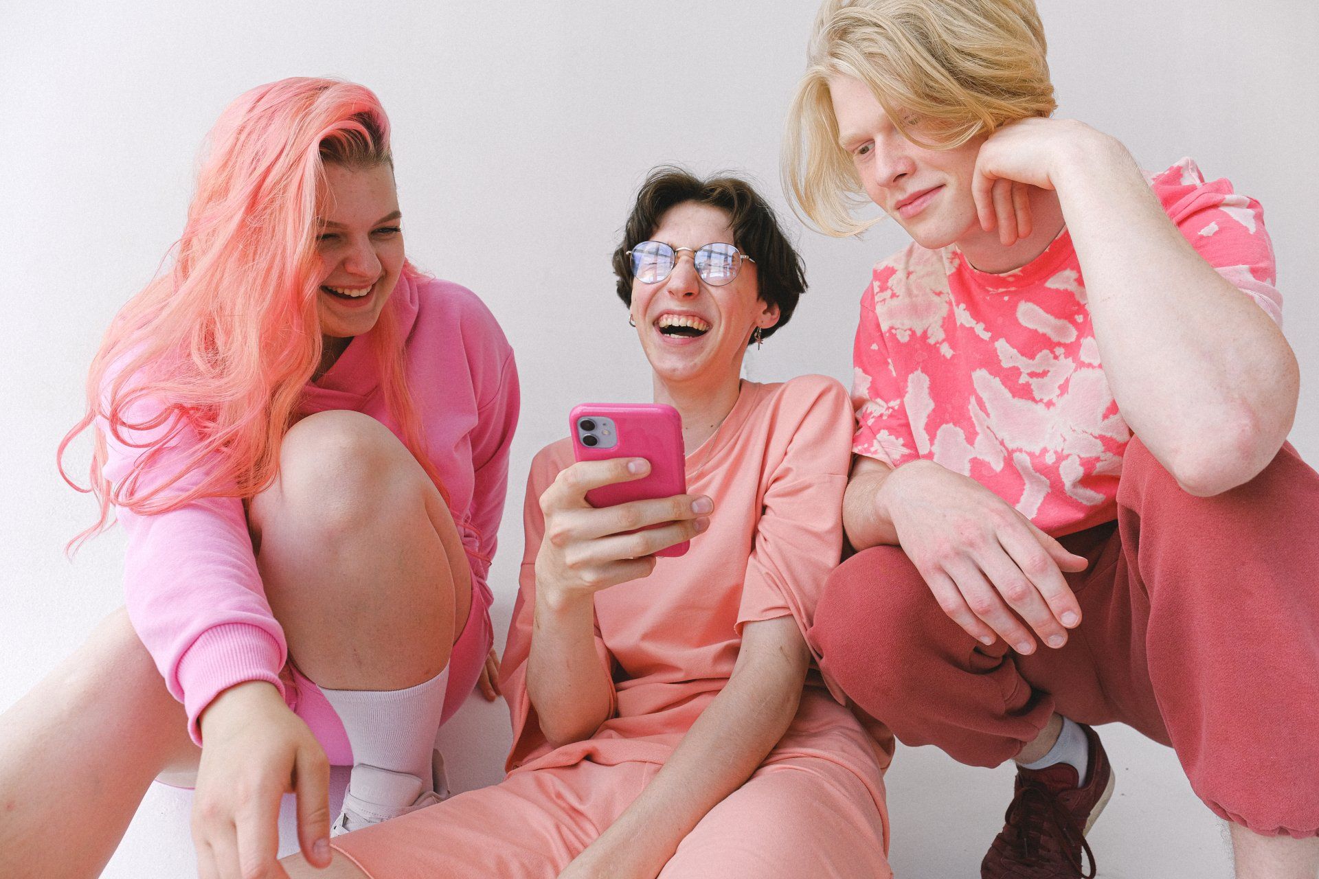 Tres jóvenes, riendo, miran un teléfono rosa. Uno tiene el pelo rosa, el otro lleva gafas. Fondo blanco, con ropa rosa.