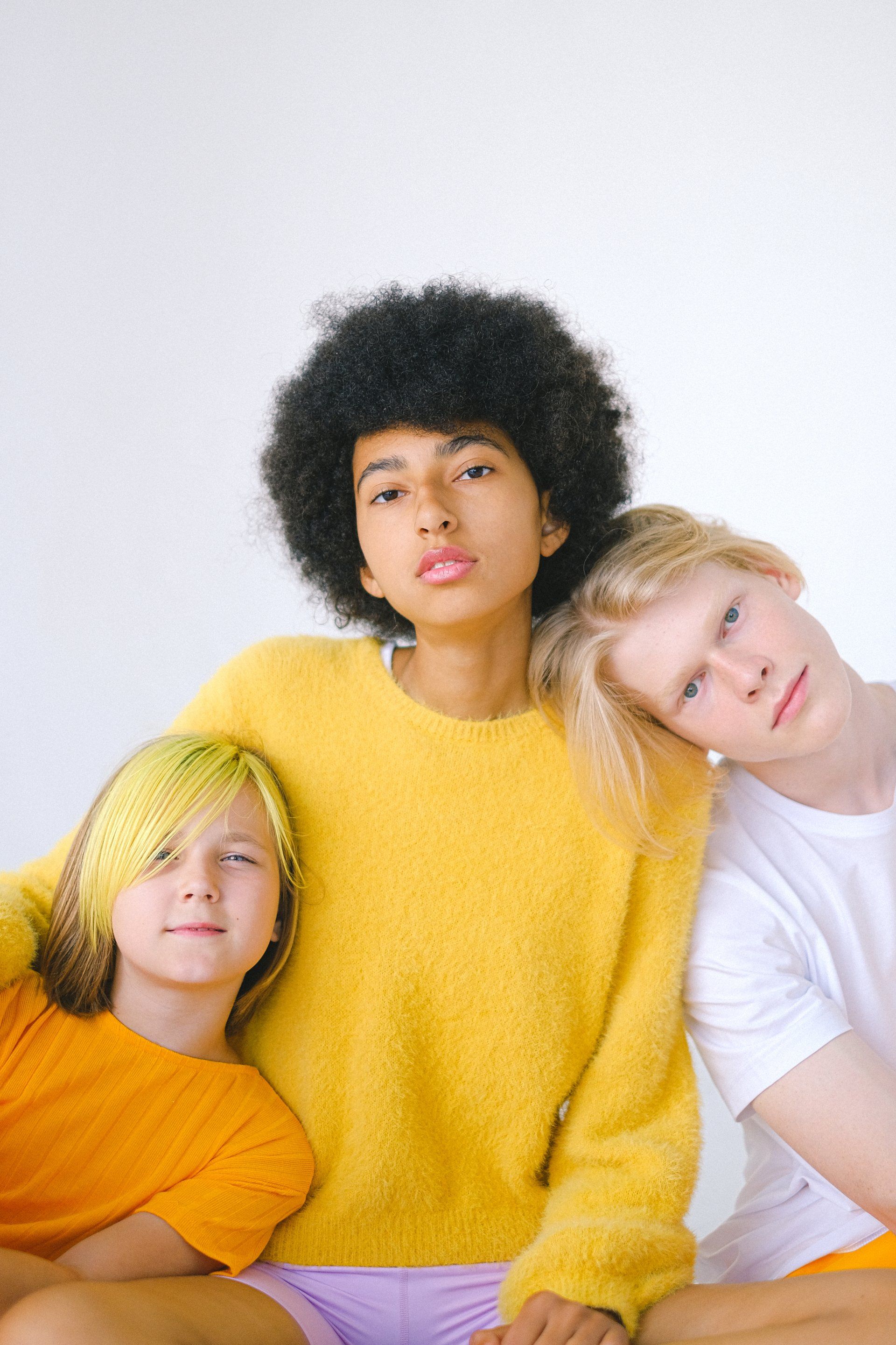 Three people leaning together against a white background, wearing yellow, orange, and white tops.