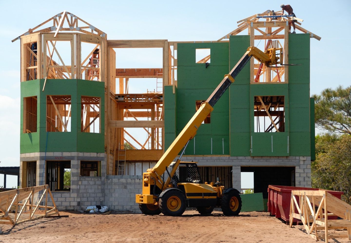 House under construction with green siding, yellow forklift extending upwards, and wooden framing.