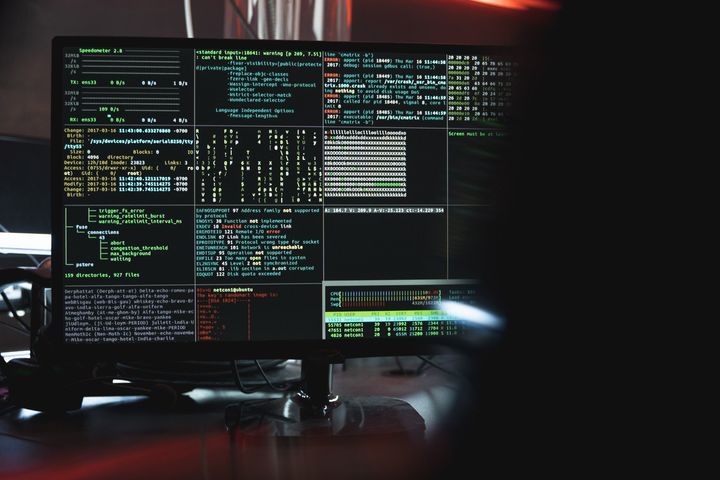 Reboot IT Hacker at a computer monitor displaying complex code in green and white, dark room, red glow.
