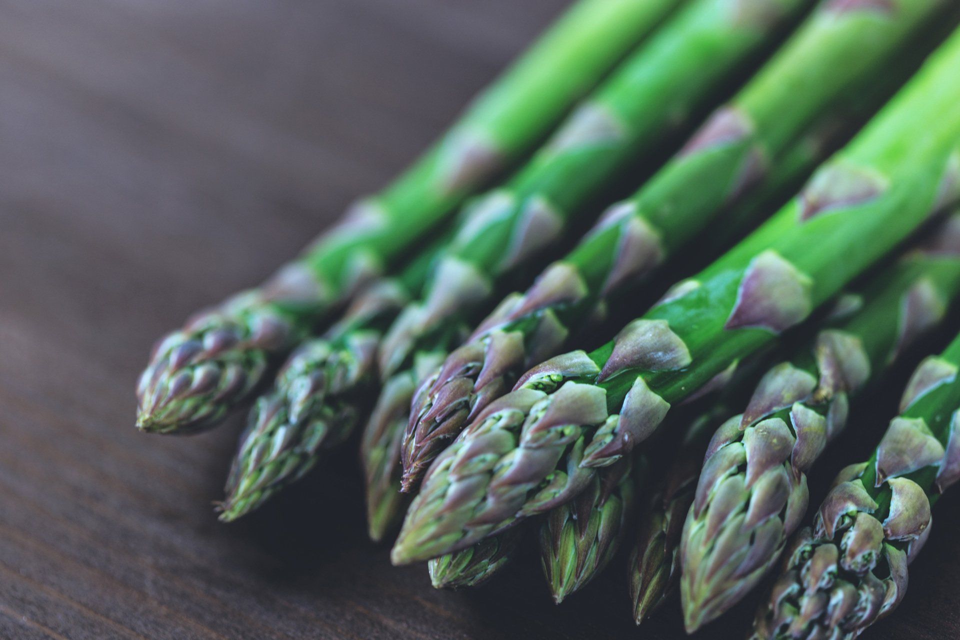 A close-up view of fresh green asparagus spears with purplish tips resting on a dark wooden surface.