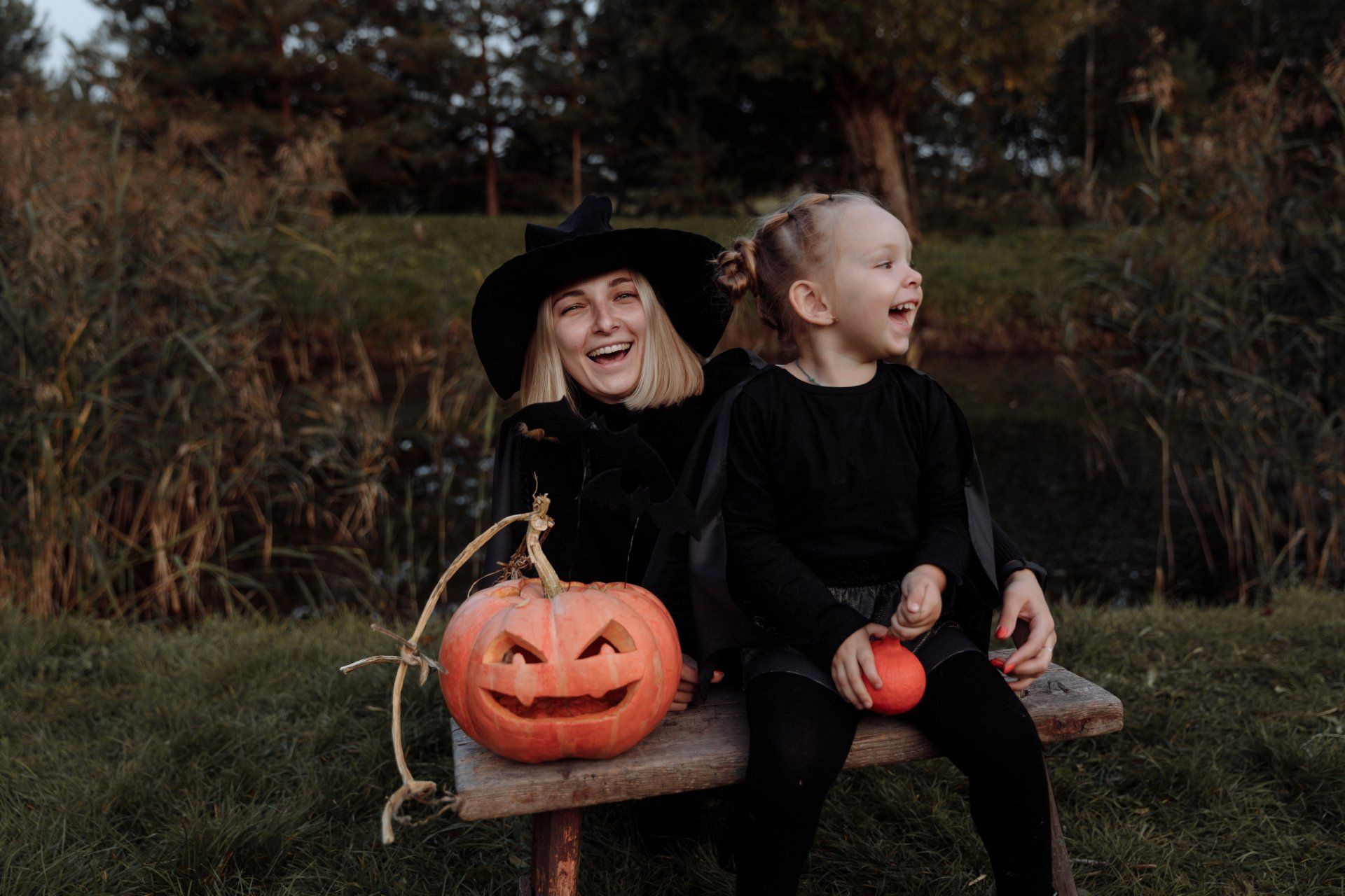 A person in a witch hat and a laughing child sit on a bench with a carved jack-o'-lantern near a pond at dusk.