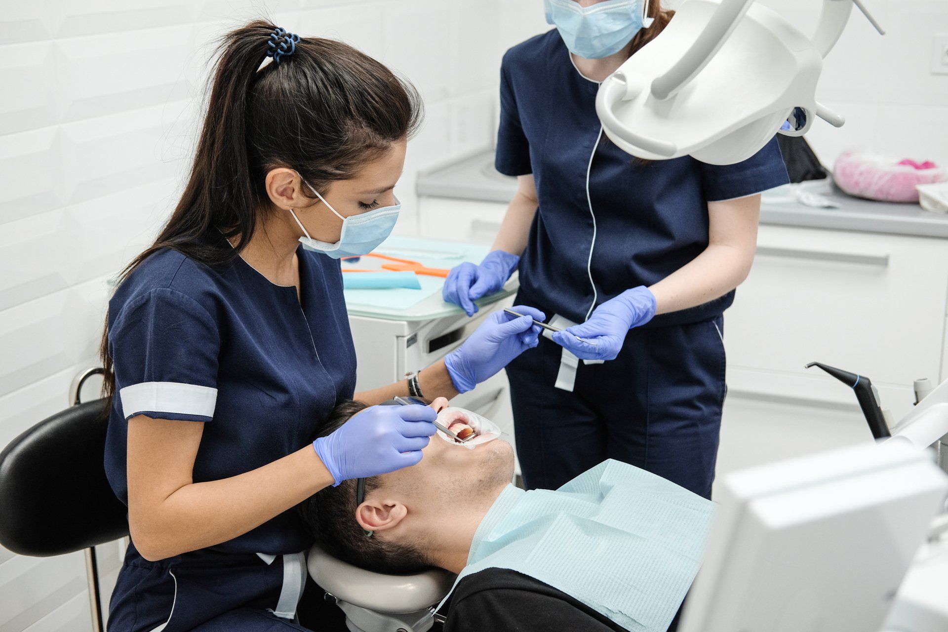 Dentist with assistant examines patient's teeth in dental office, wearing masks and blue scrubs.