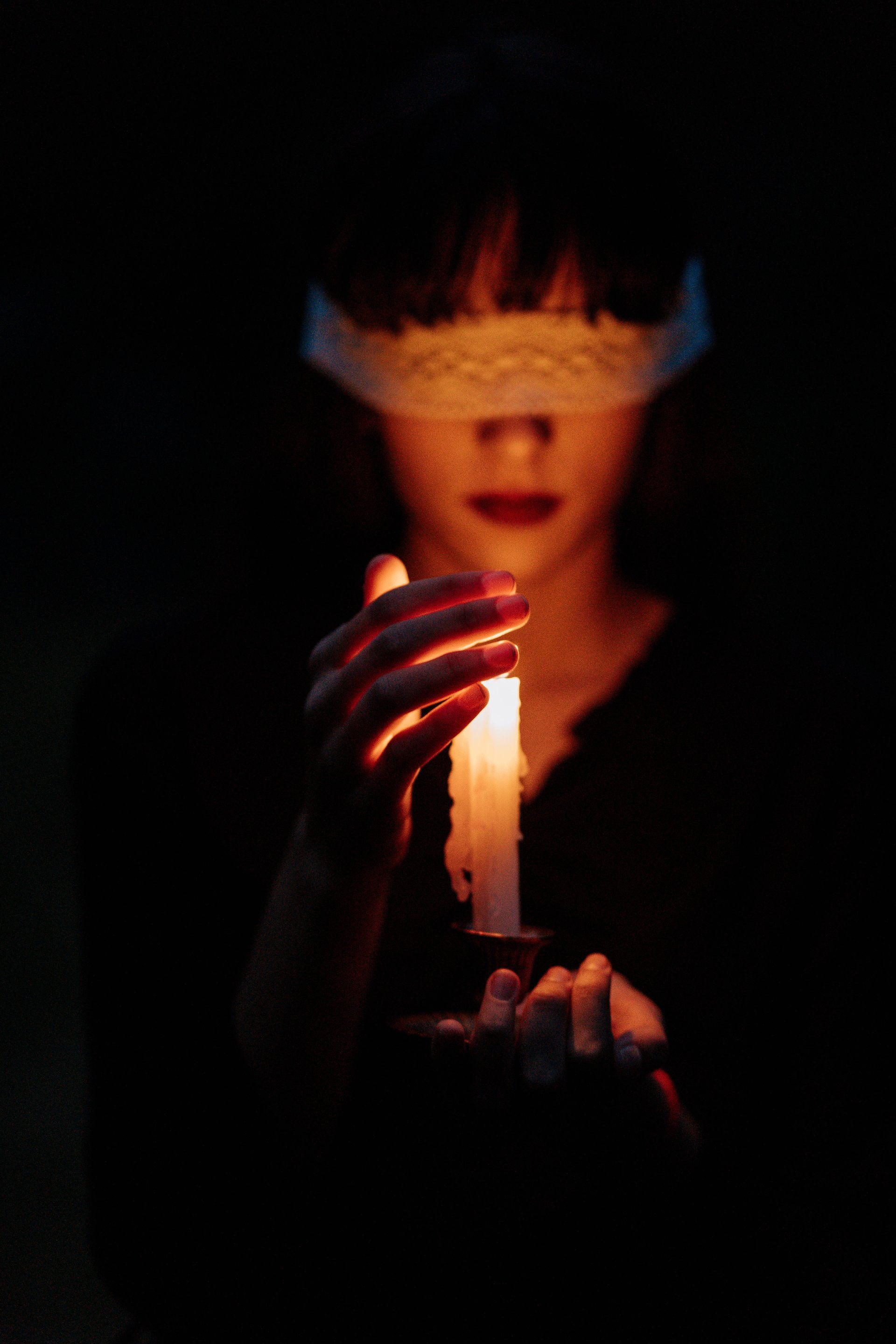 A person with a white lace blindfold holds a lit, melting candle in the dark, casting a warm glow on their face.