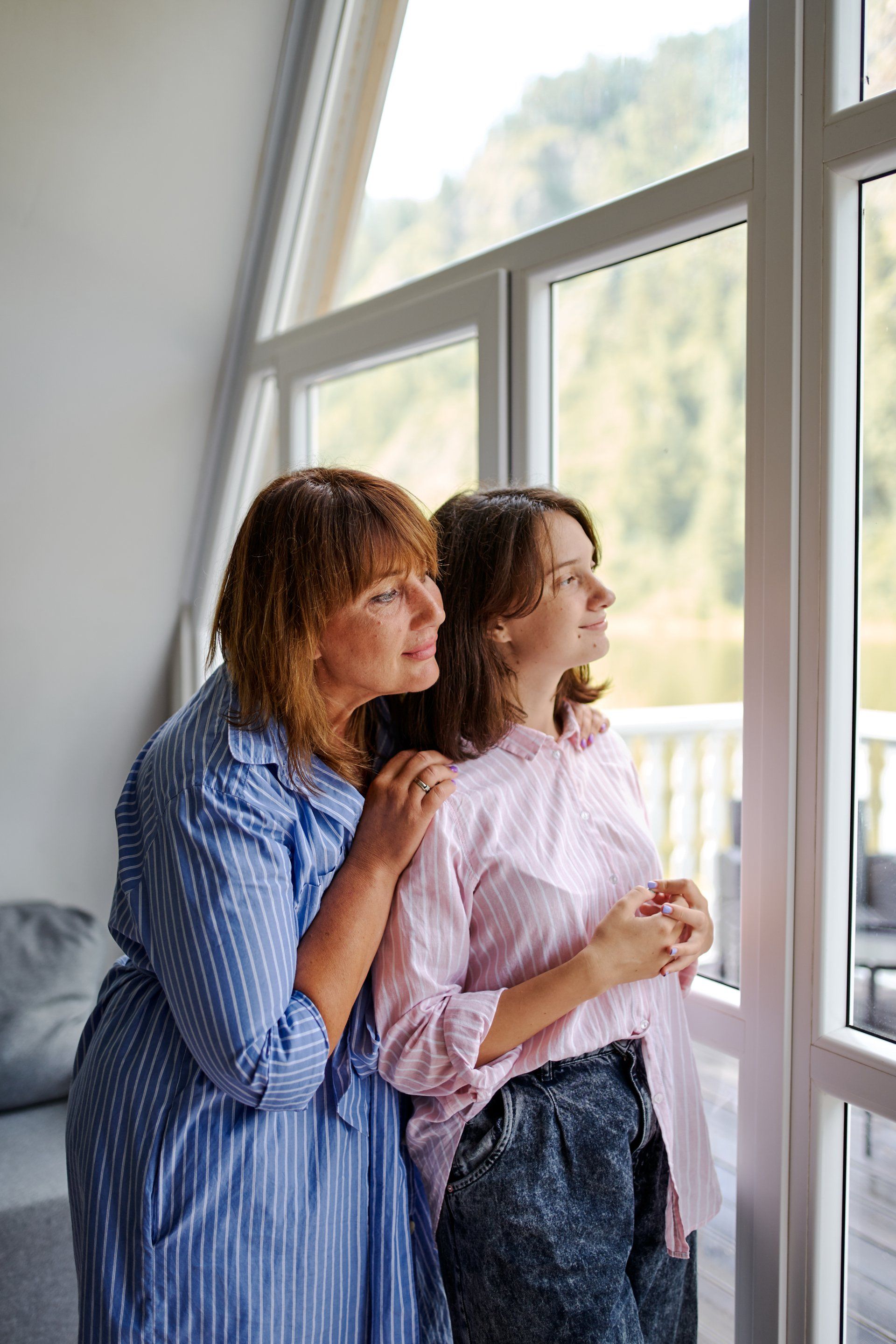 Two women are standing next to each other in front of a window looking out.