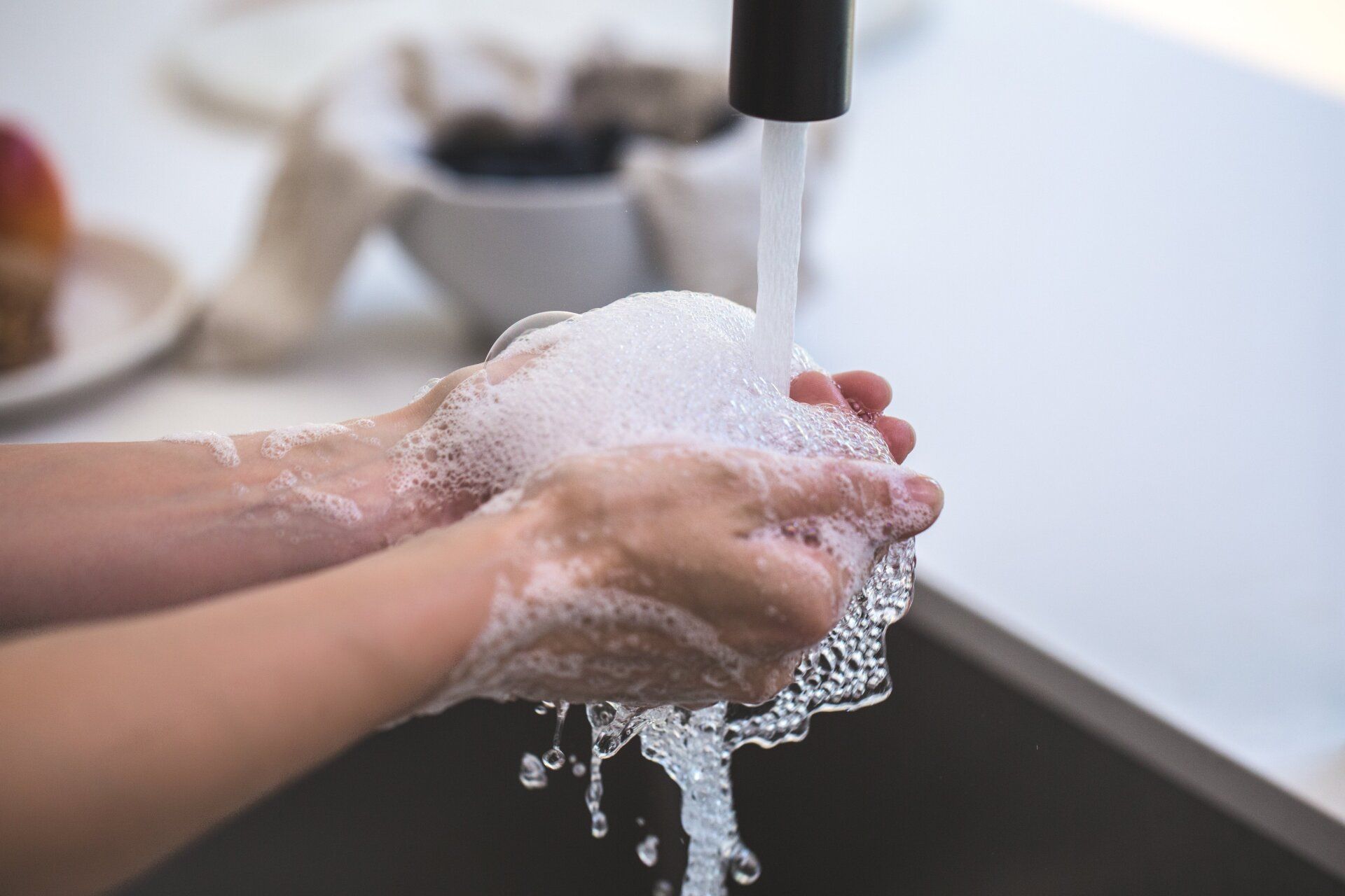 Hands lathered in soap bubbles being rinsed under a running kitchen faucet.