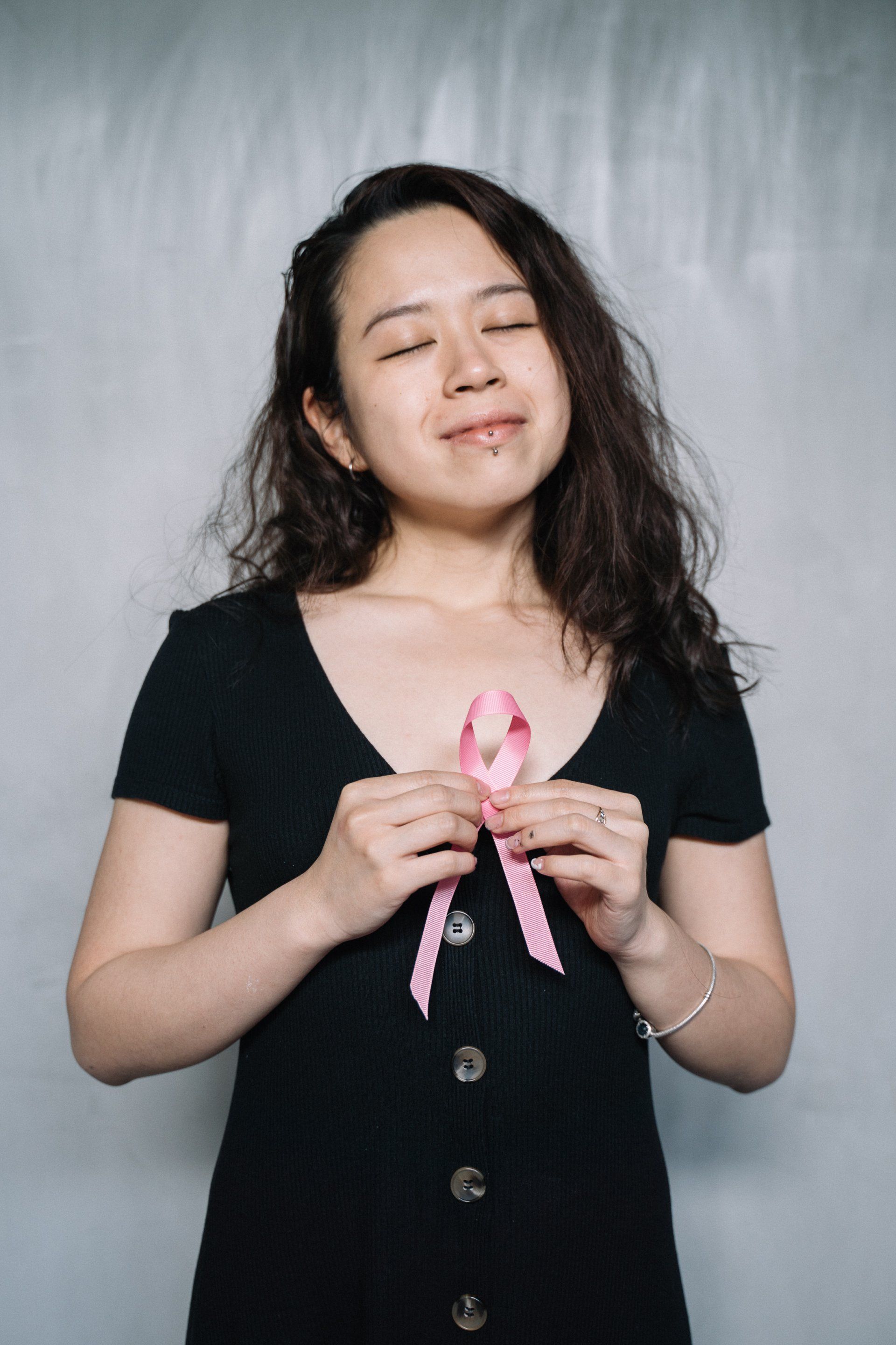 Woman holding a pink ribbon with a hopeful expression. Black dress, gray background.