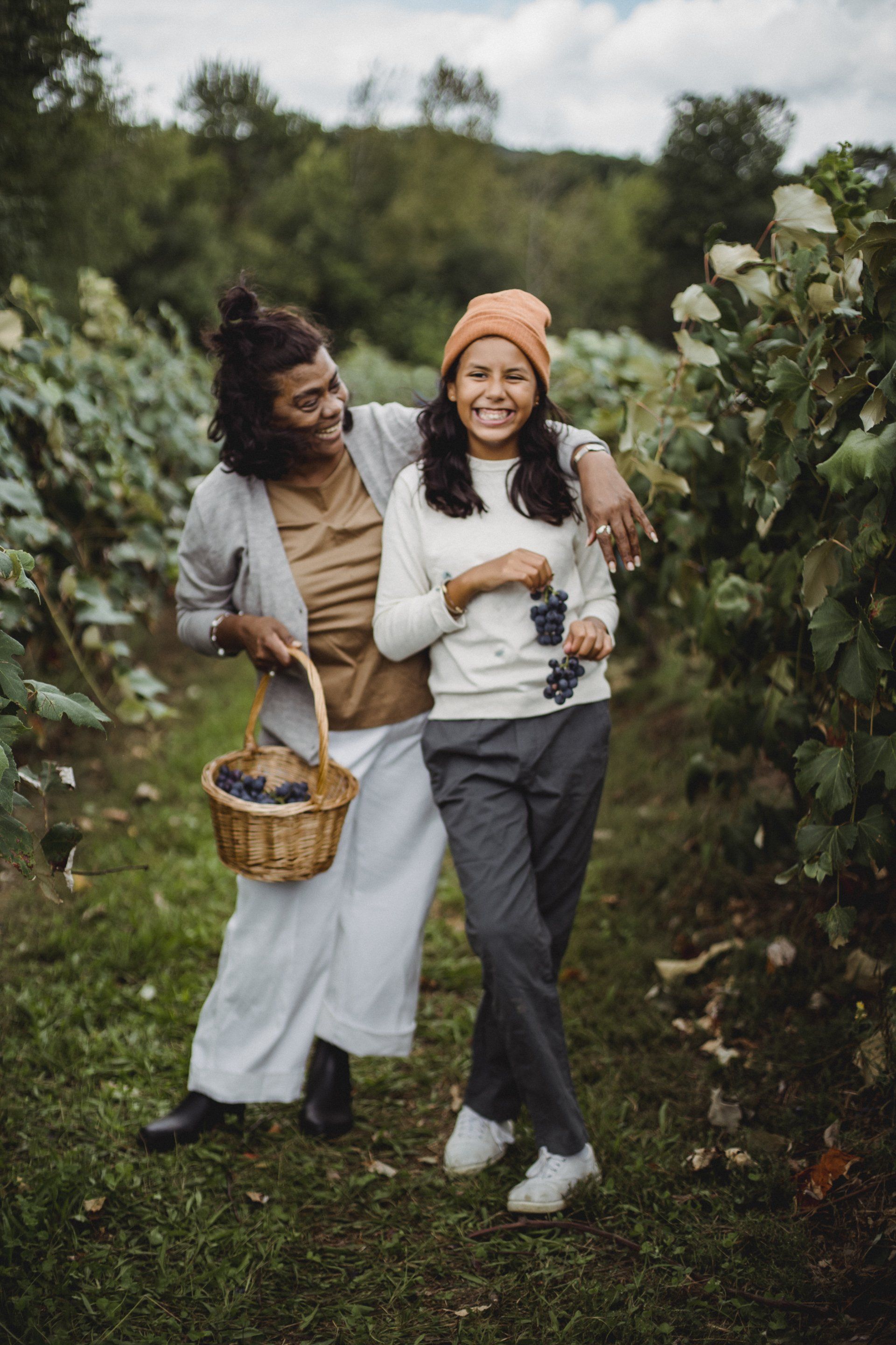 Two women are standing next to each other in a vineyard holding baskets of grapes.