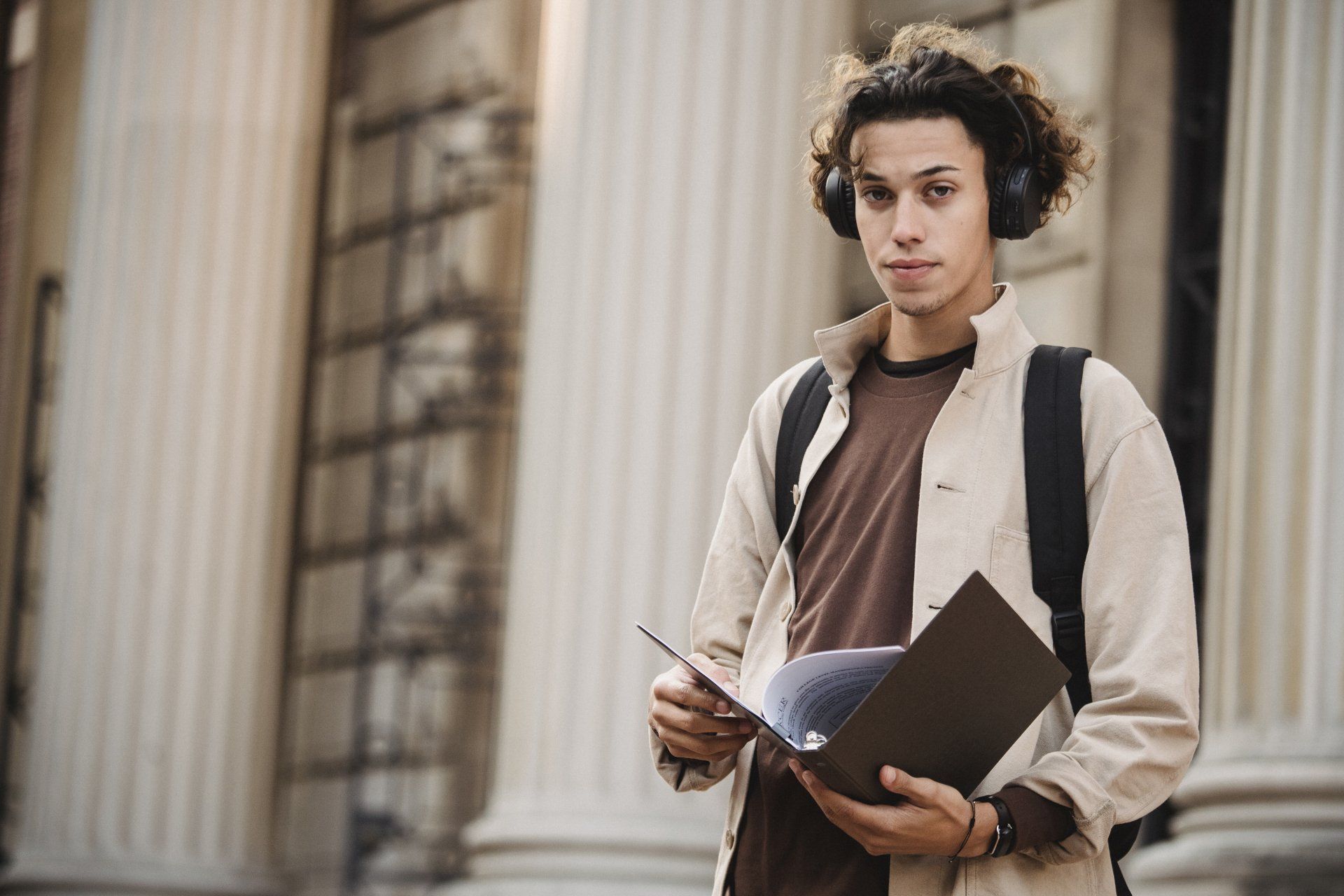 A young man wearing headphones and holding a book is standing in front of a building.