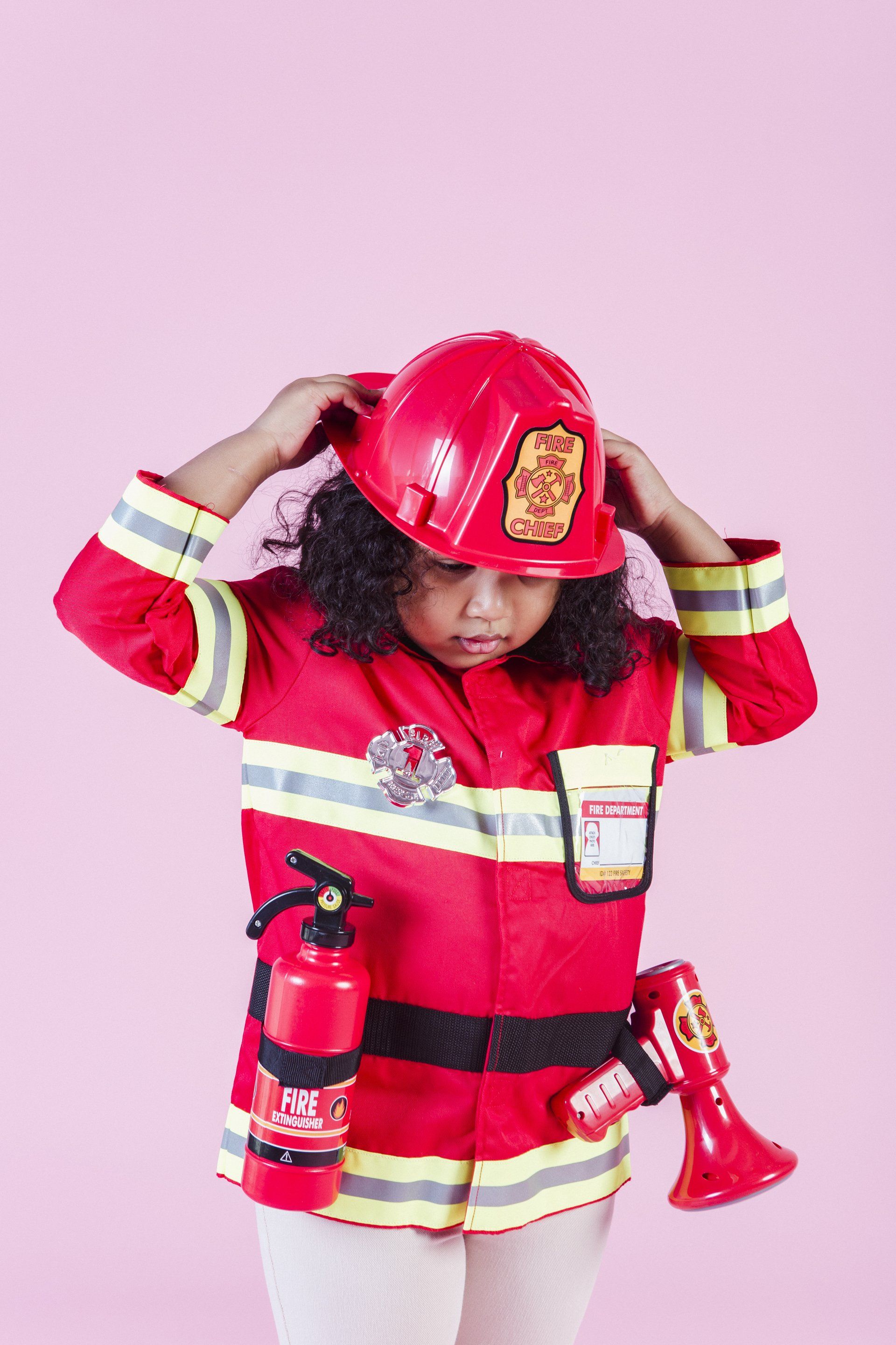 A little girl in a fireman costume is putting on her helmet.