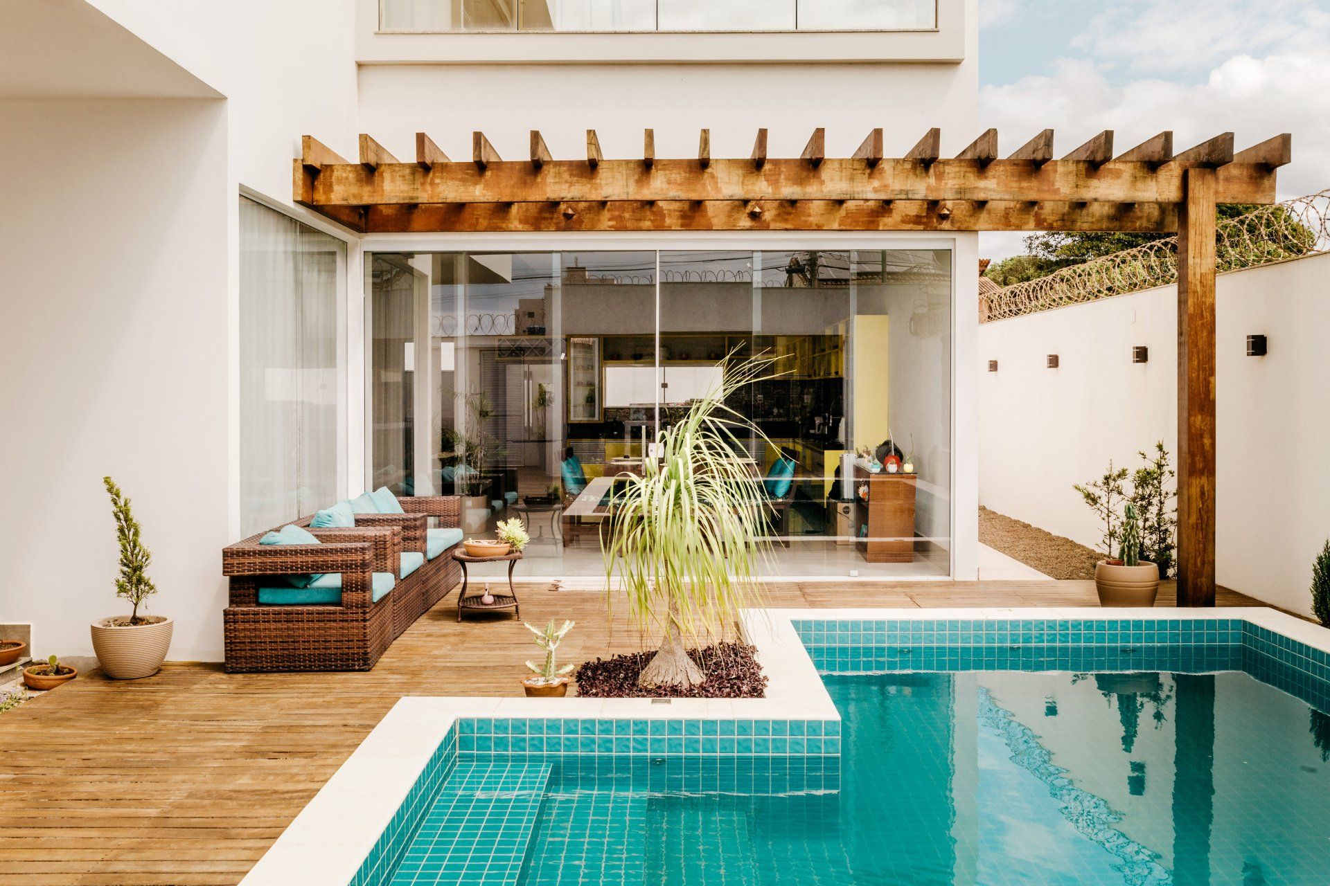 Modern patio with turquoise pool, wooden deck, pergola, and glass doors opening to a home interior