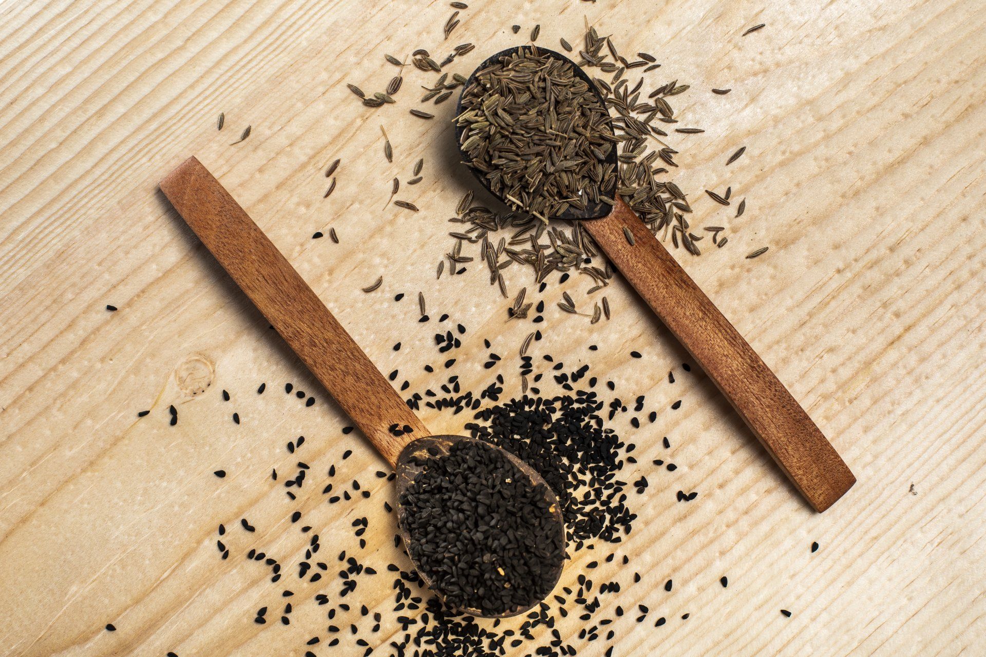 Two wooden spoons filled with black and brown seeds on a light wooden surface.