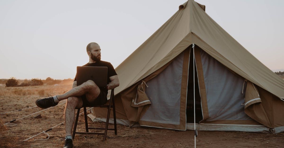 A man is sitting in a chair in front of a tent using a laptop.