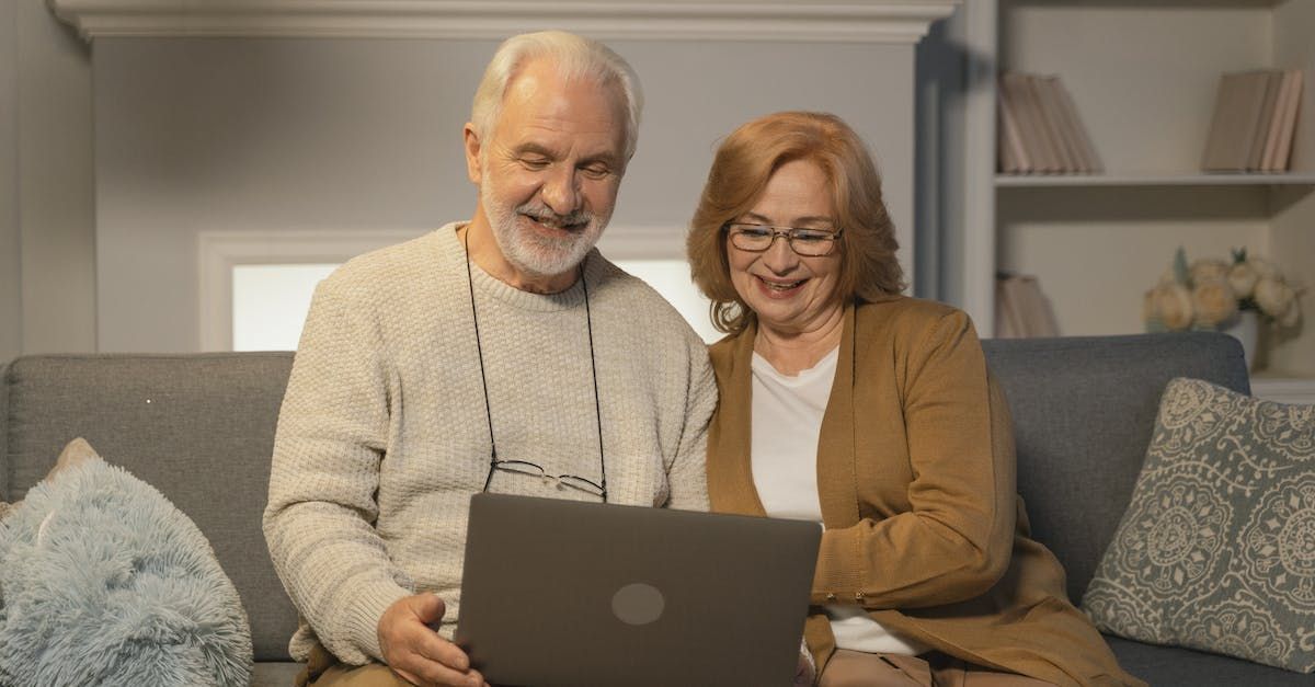 Twee mensen zitten glimlachend op een bank in de woonkamer terwijl ze samen naar een laptop kijken.