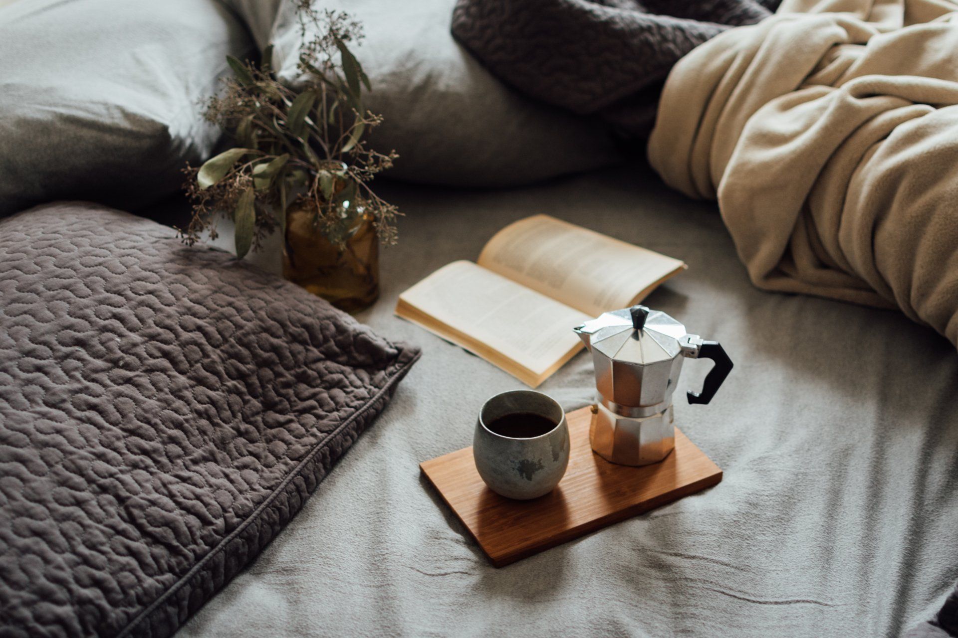 A cozy bed setting with a book, a moka pot, and a cup of coffee on a wooden tray, next to a vase with dried branches.