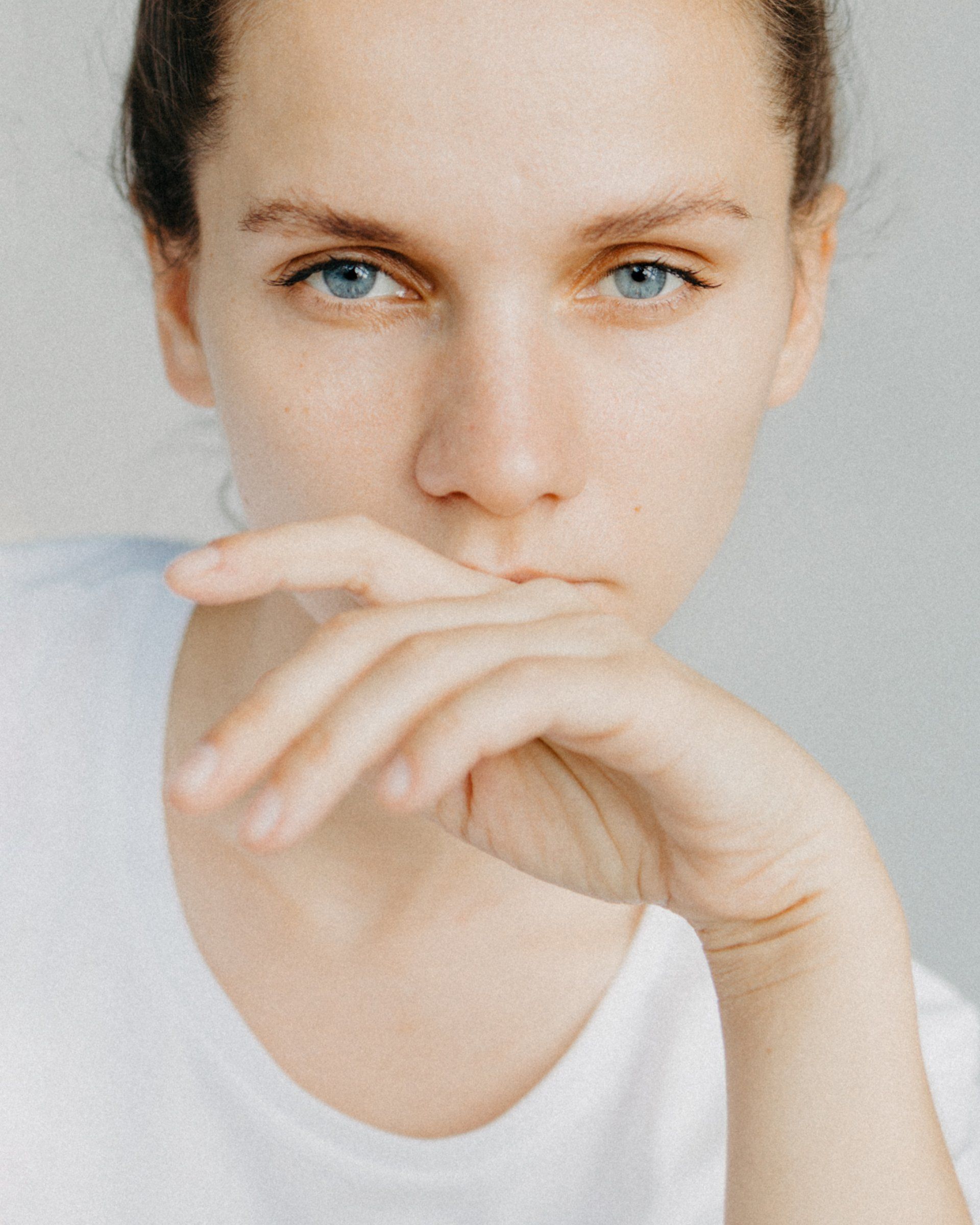 Woman with blue eyes, hand near her mouth, wearing a white t-shirt, looking at the camera.