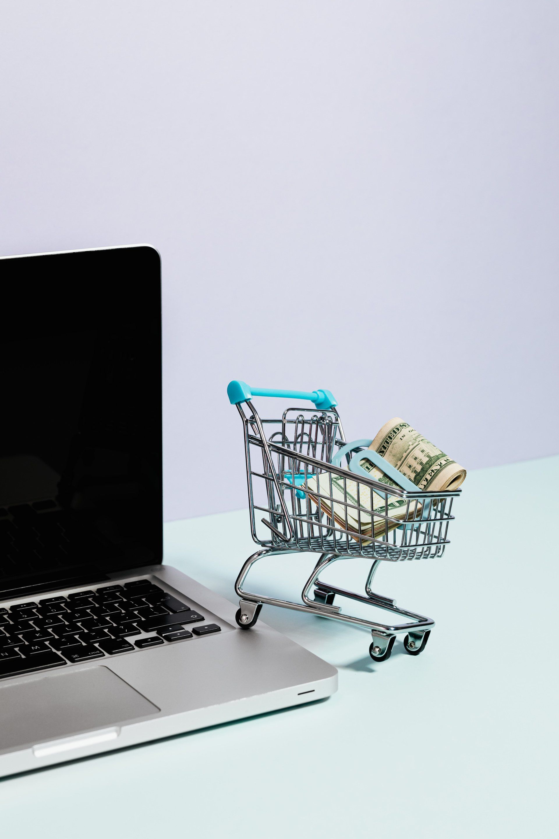 A shopping cart filled with money sits next to a laptop