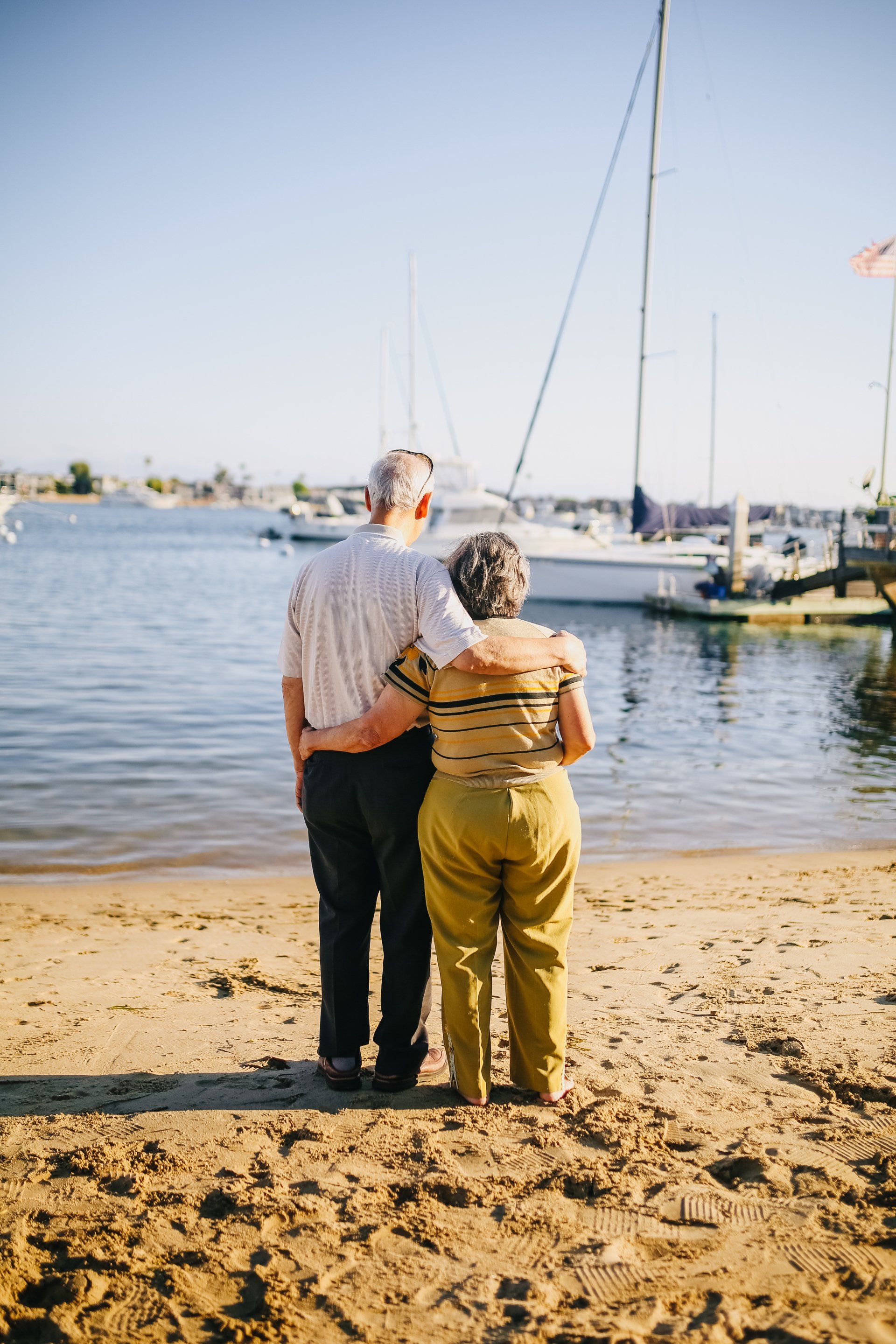 Two people stand arm in arm on a sandy shore, facing a calm marina with sailboats.